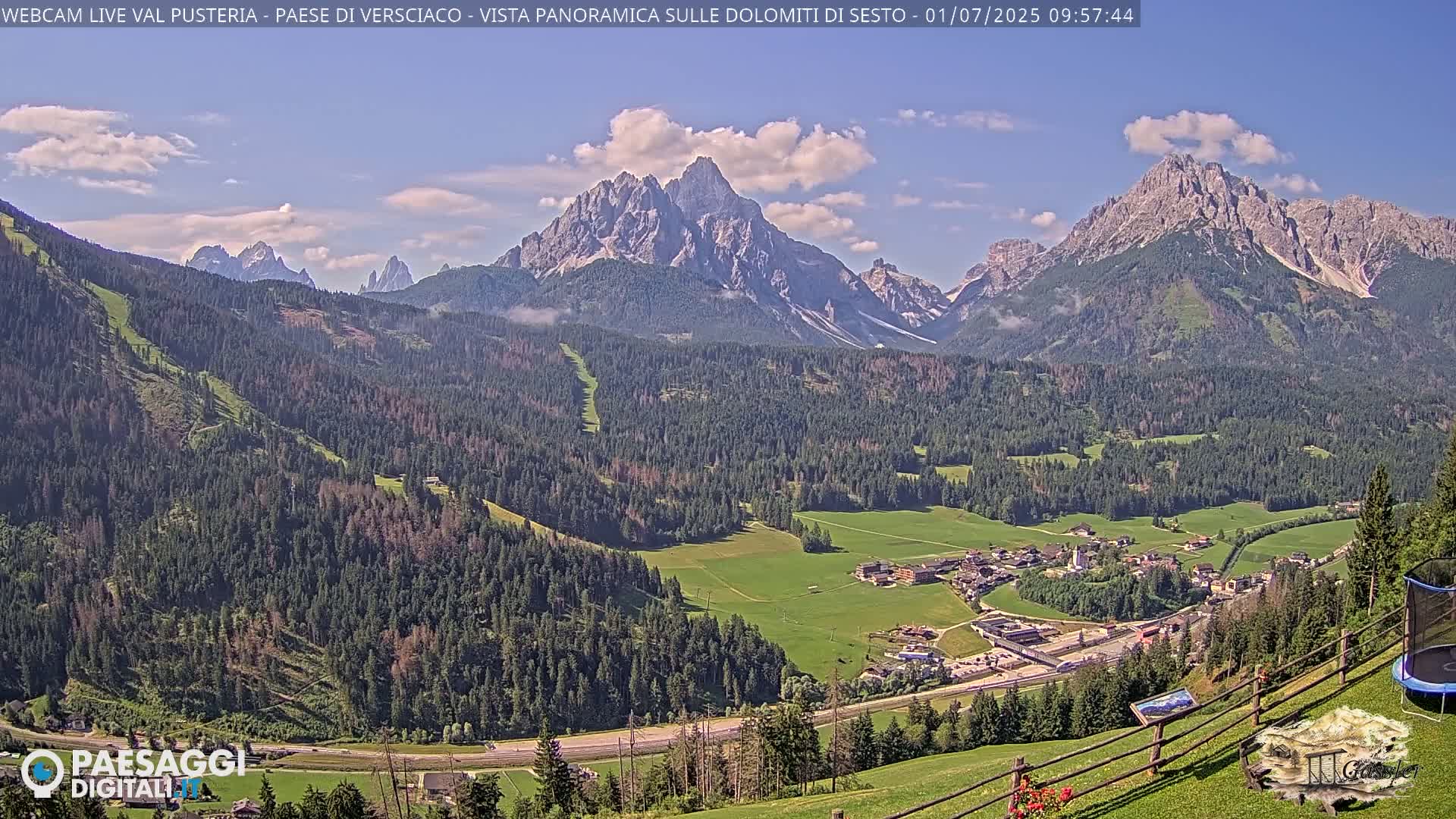 A panoramic view shows a valley with a village nestled amongst evergreen forests, overlooked by imposing, rocky mountains under a partly cloudy sky.
