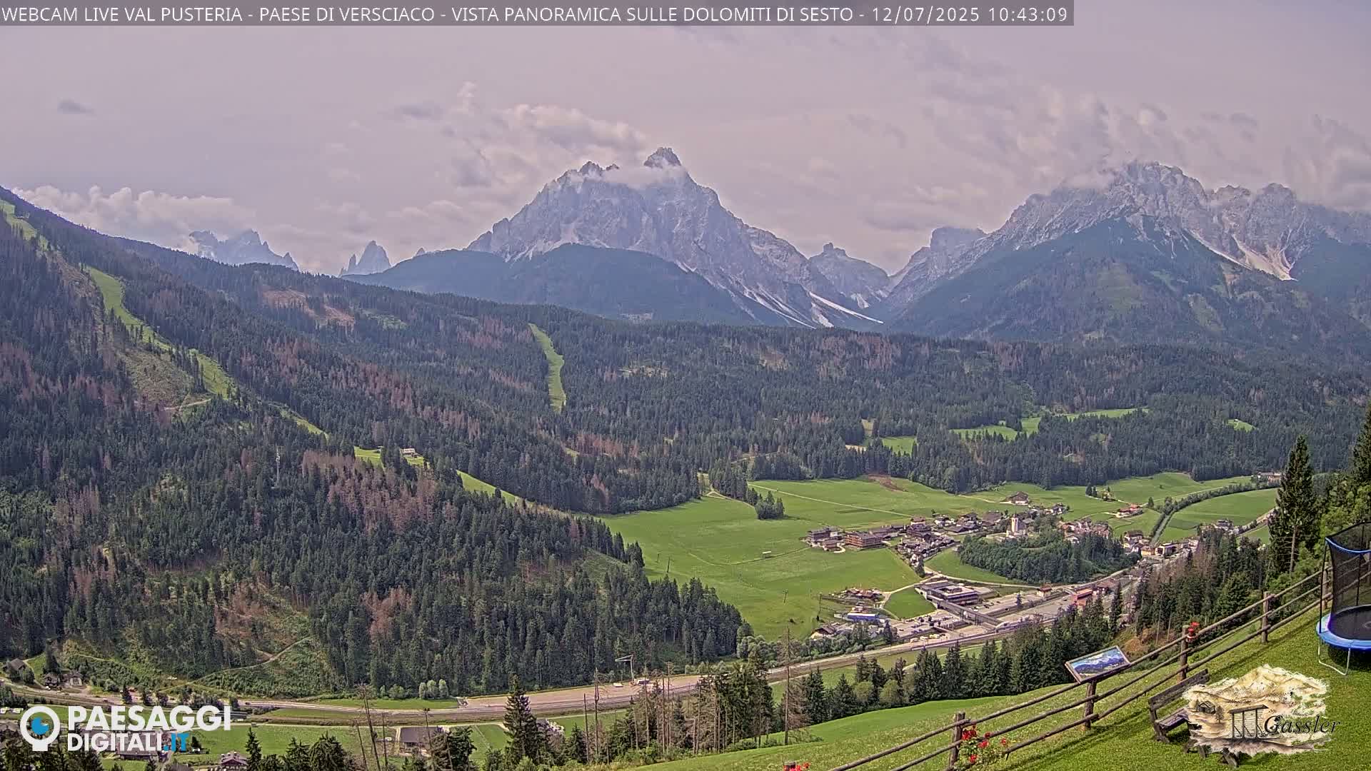 A panoramic view shows a valley with a village nestled amongst evergreen forests, overlooked by imposing, rocky mountains under a partly cloudy sky.