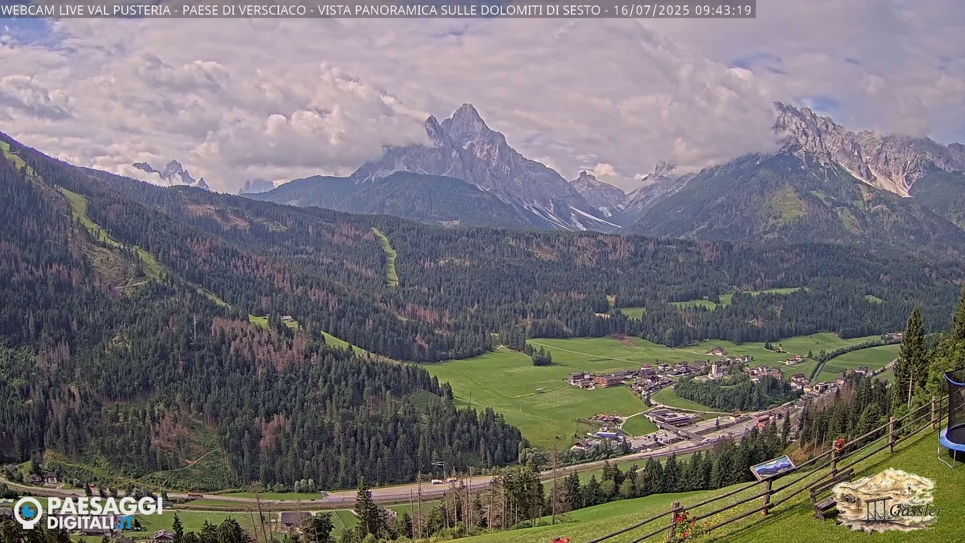 A partly cloudy mountain valley scene shows a village nestled amongst green fields and evergreen forests, with a prominent, snow-dusted peak in the background.