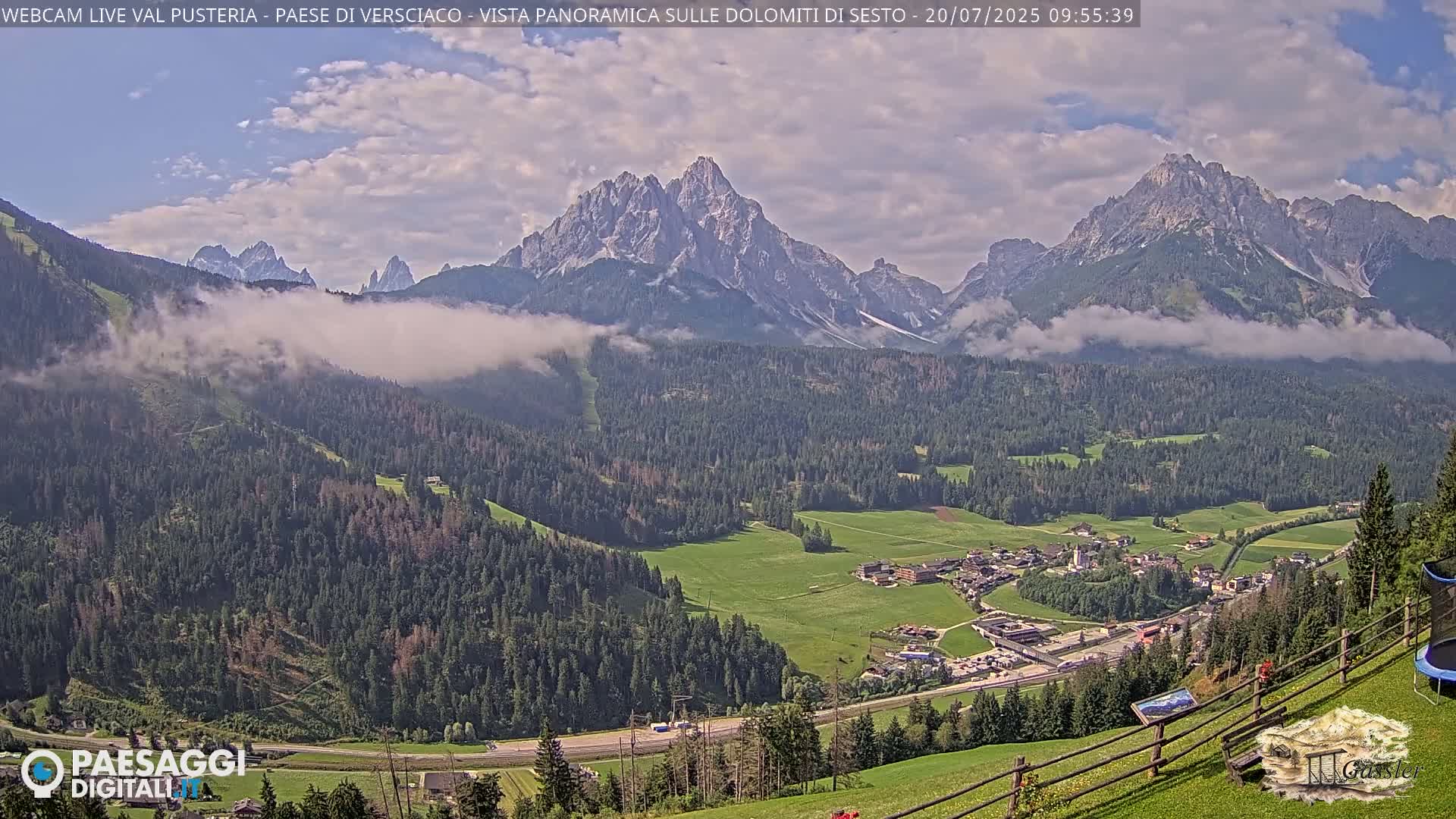 A valley town nestled among lush green forests and partially cloudy skies, with a majestic mountain range in the background.
