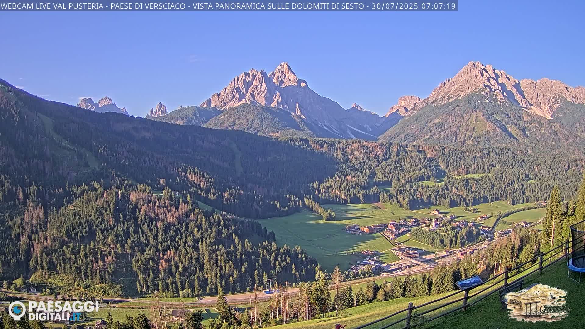 A valley town nestled among lush green forests and partially cloudy skies, with a majestic mountain range in the background.