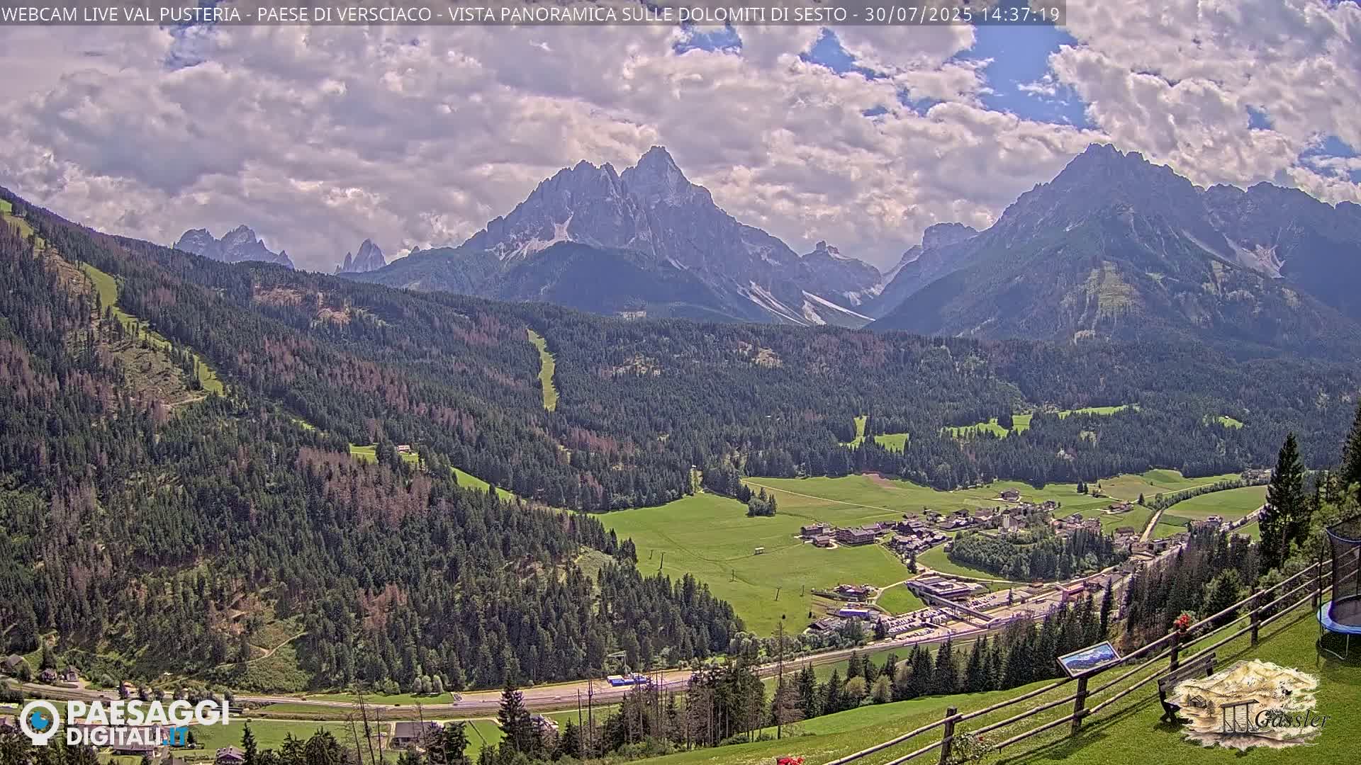 A valley town nestled among lush green forests and partially cloudy skies, with a majestic mountain range in the background.