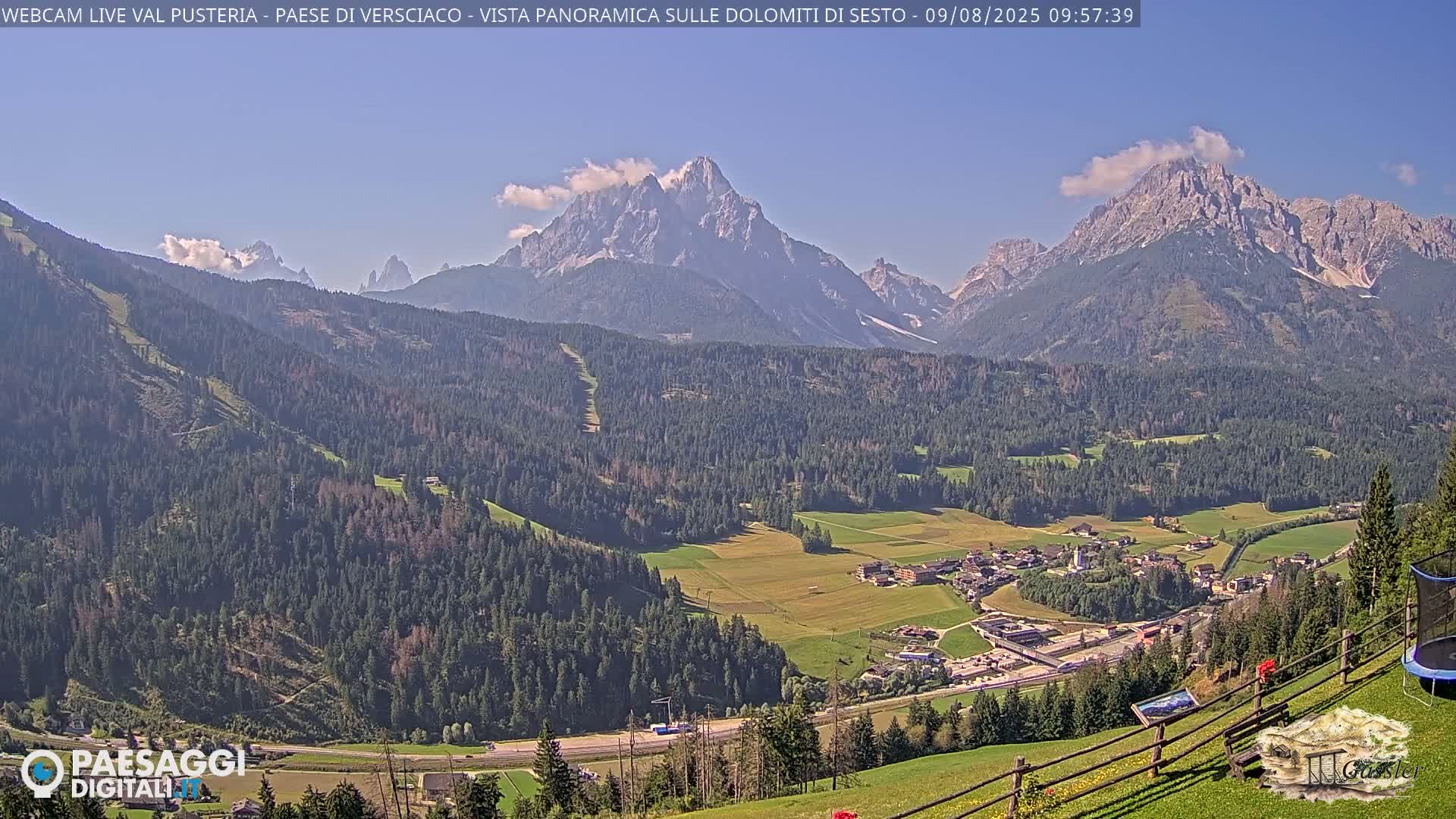 A panoramic view shows a valley town nestled amongst green hills and forests, with towering, snow-capped mountains in the background under a clear blue sky.