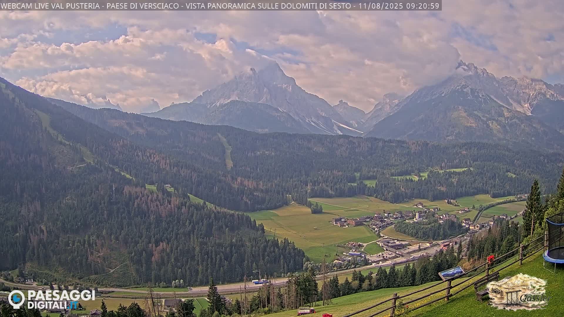 A partly cloudy day reveals a panoramic view of a valley town nestled among verdant forests and towering, snow-capped mountains.