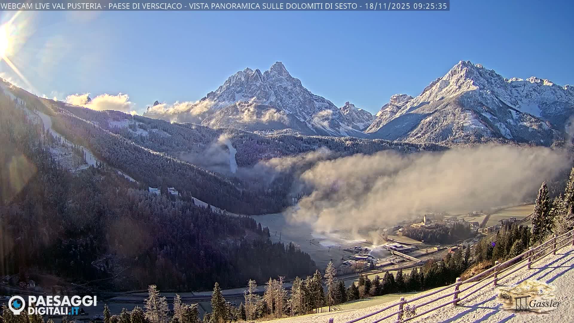 The image shows a panoramic view of a snow-covered mountain valley with a town nestled below, bathed in bright morning sunlight under a clear blue sky, with mist or low clouds lingering in the mid-ground.