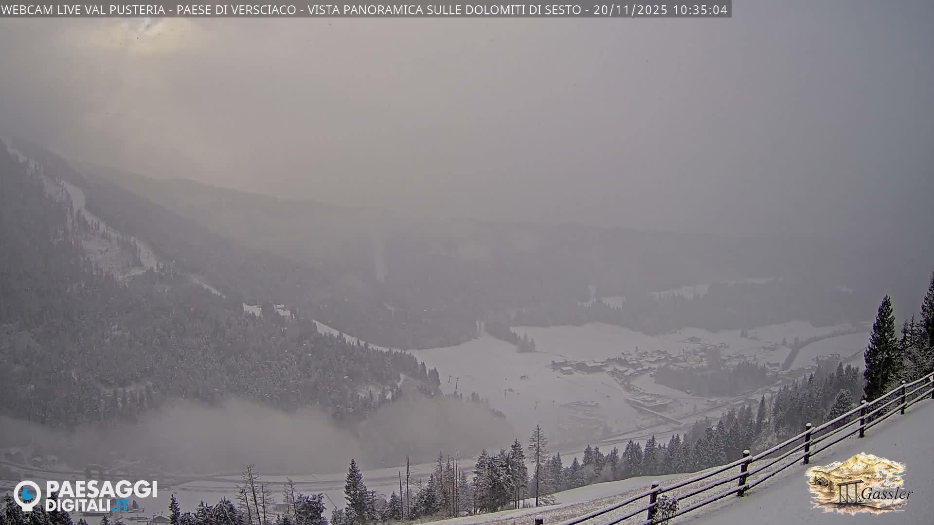A wide panoramic view captures a snow-covered mountain valley with forested slopes, a distant village, and ski trails, all under an overcast and hazy winter sky.