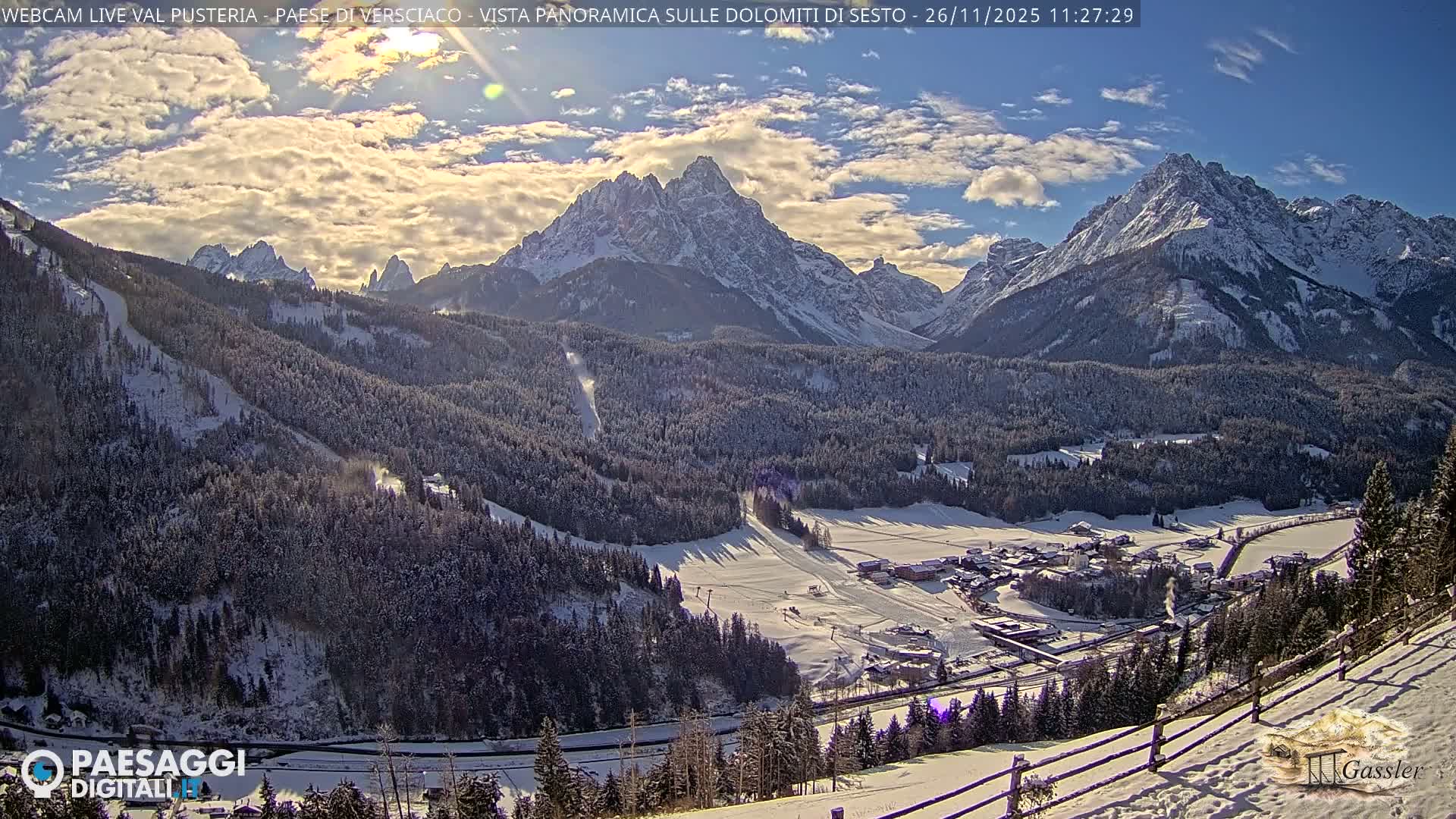 A wide panoramic view captures a snow-covered mountain valley with forested slopes, a distant village, and ski trails, all under an overcast and hazy winter sky.