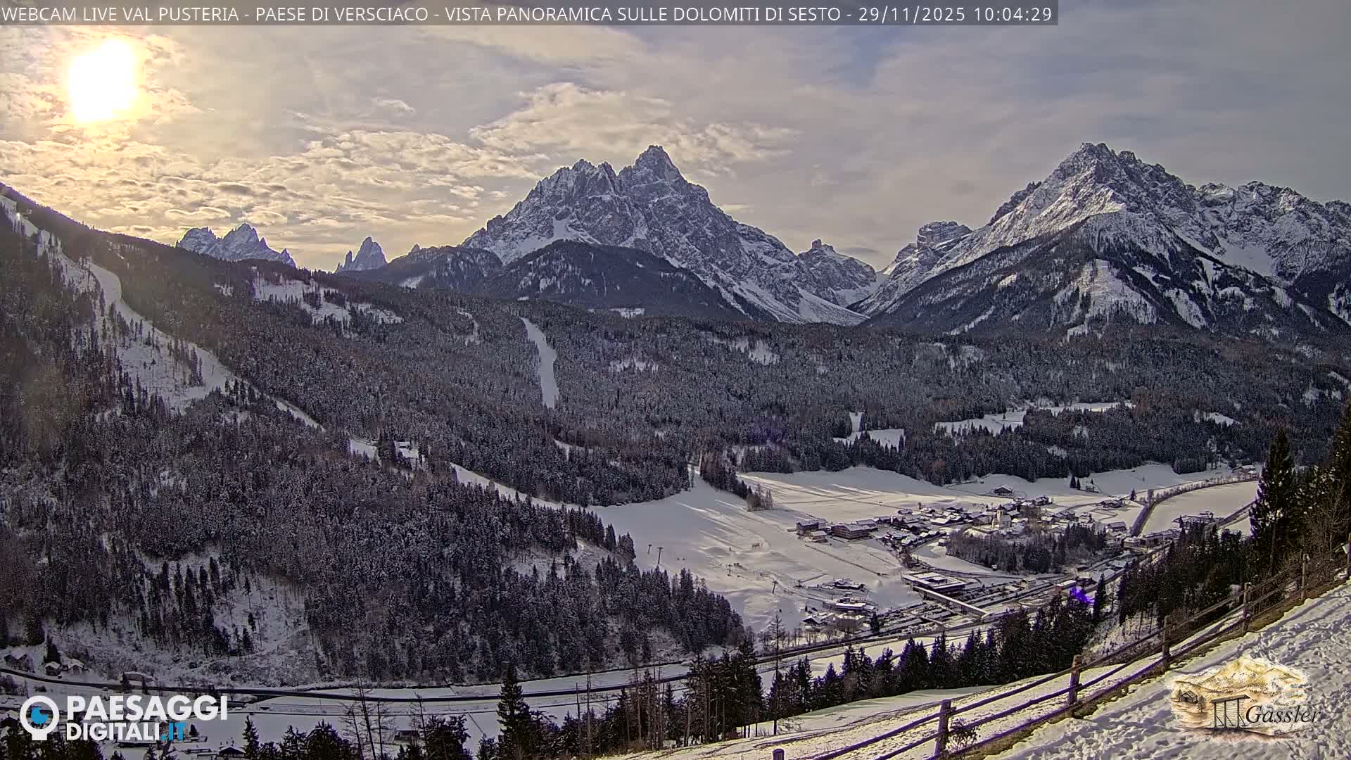 A panoramic winter landscape reveals majestic snow-capped mountains towering over a valley filled with snow-covered coniferous forests, a small village, and a visible ski slope, all bathed in bright sunlight under a partly cloudy sky.
