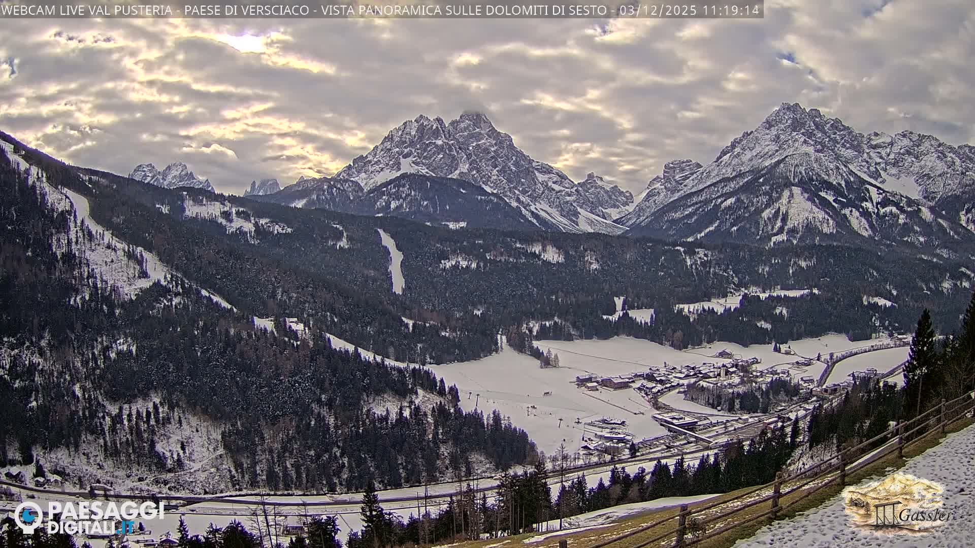 A panoramic winter landscape reveals a snow-covered valley with a village and dark evergreen forests, bordered by towering, snow-capped mountains beneath a heavily clouded sky.