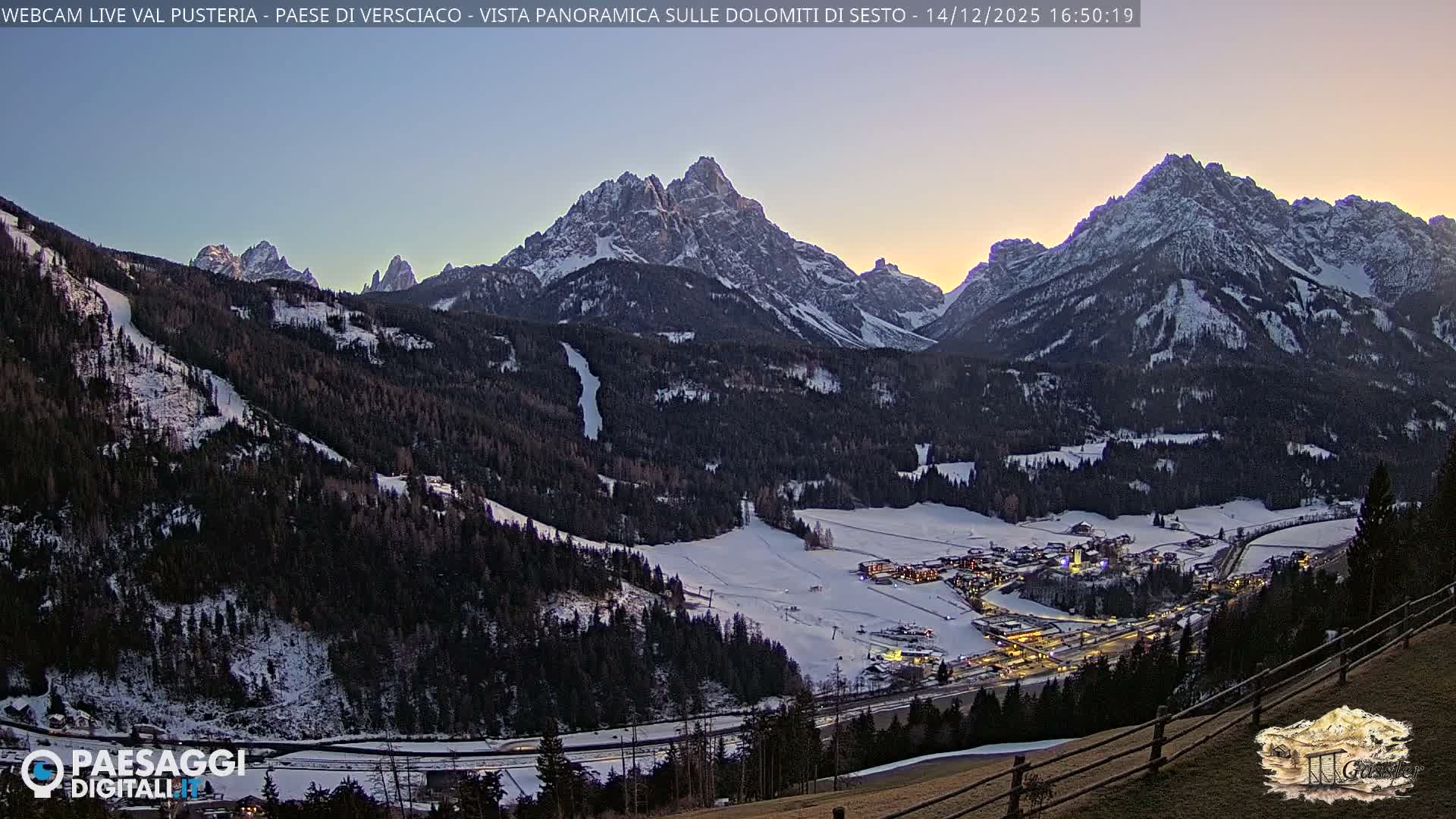 A panoramic winter landscape reveals a snow-covered valley with a village and dark evergreen forests, bordered by towering, snow-capped mountains beneath a heavily clouded sky.