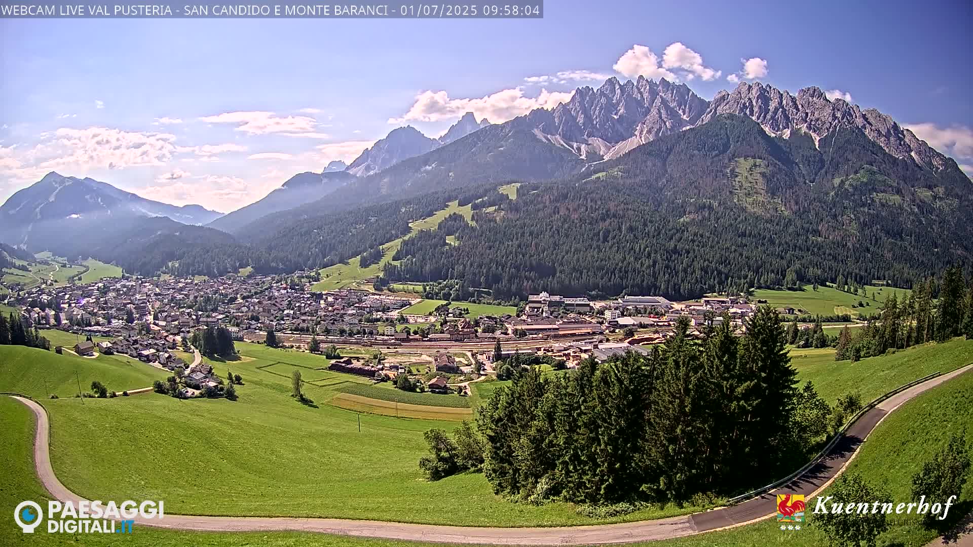 A sunny day reveals a mountain village nestled in a valley, with a majestic mountain range rising in the background.
