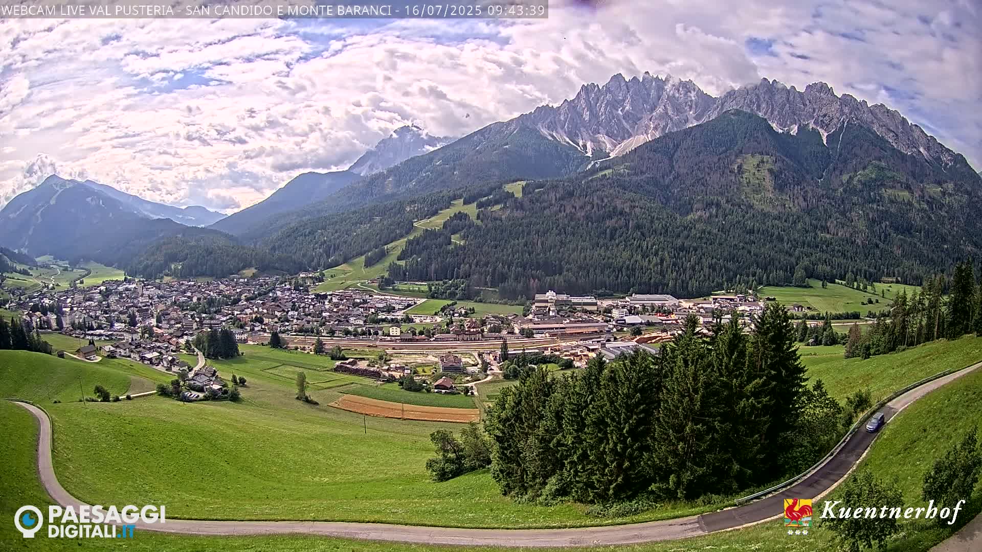 A partly cloudy day reveals a mountainous landscape, featuring a town nestled in a valley below a large, jagged mountain range.
