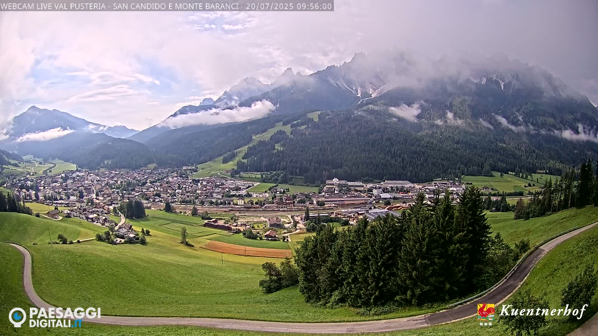 A partly cloudy day reveals a valley town nestled among green hills and mountains partially obscured by mist.