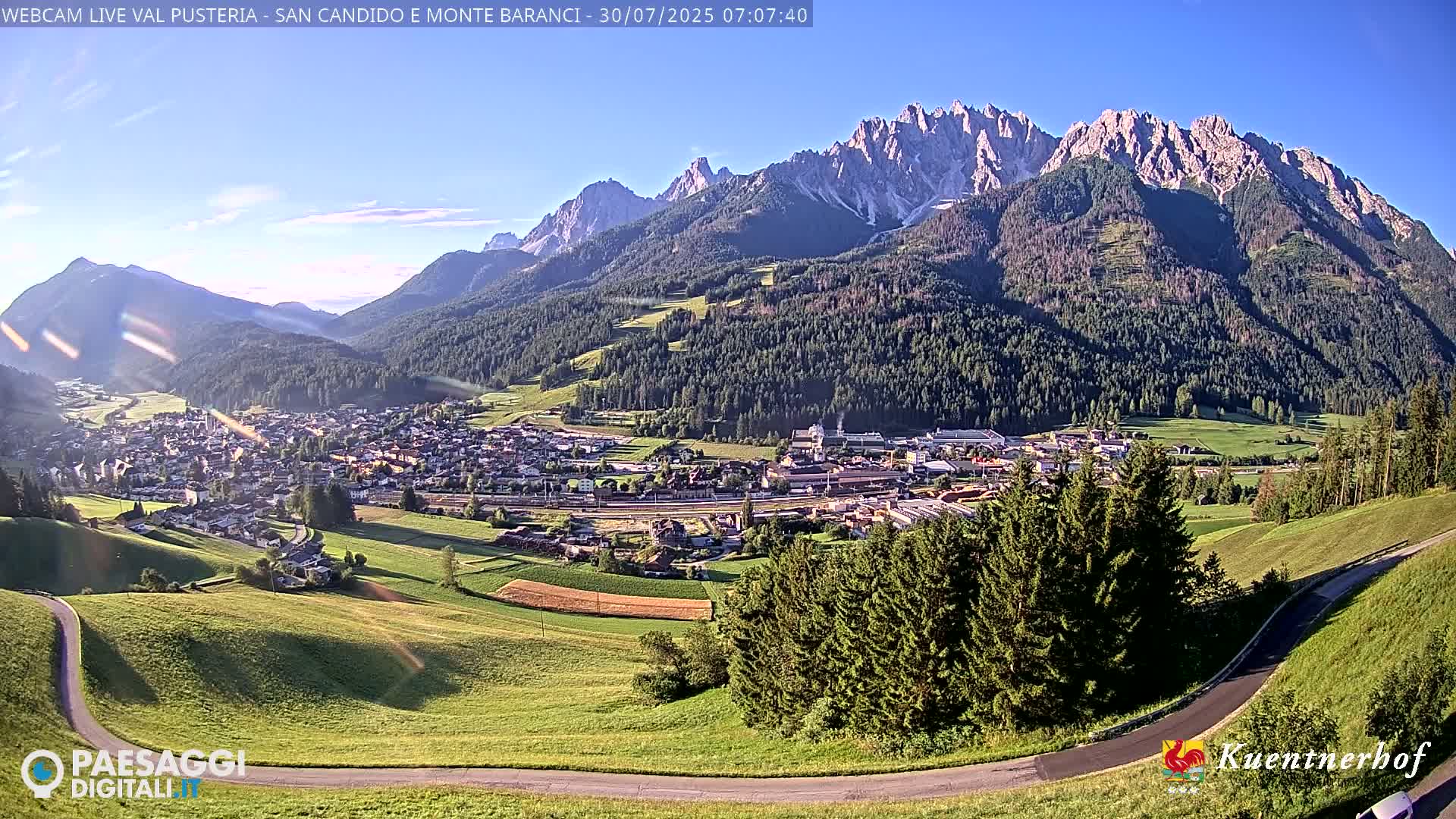 A partly cloudy day reveals a valley town nestled among green hills and mountains partially obscured by mist.