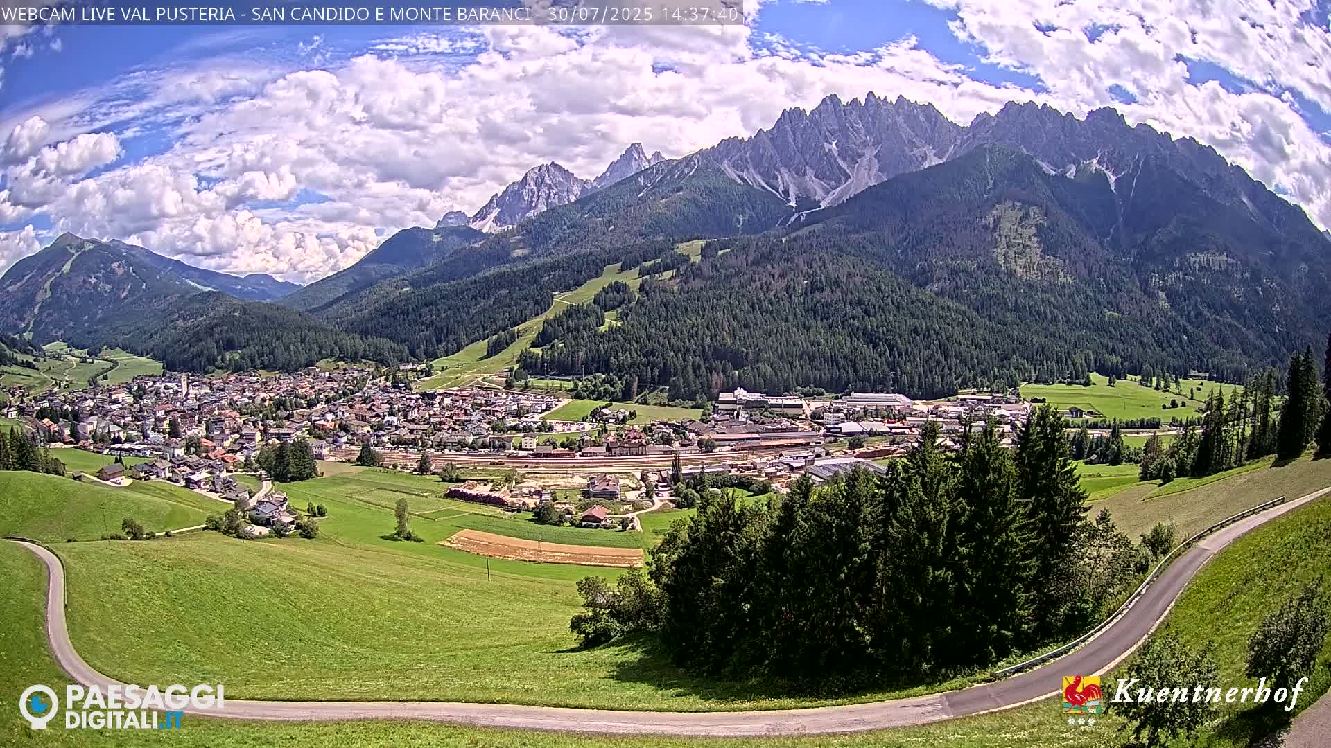 A partly cloudy day reveals a valley town nestled among green hills and mountains partially obscured by mist.