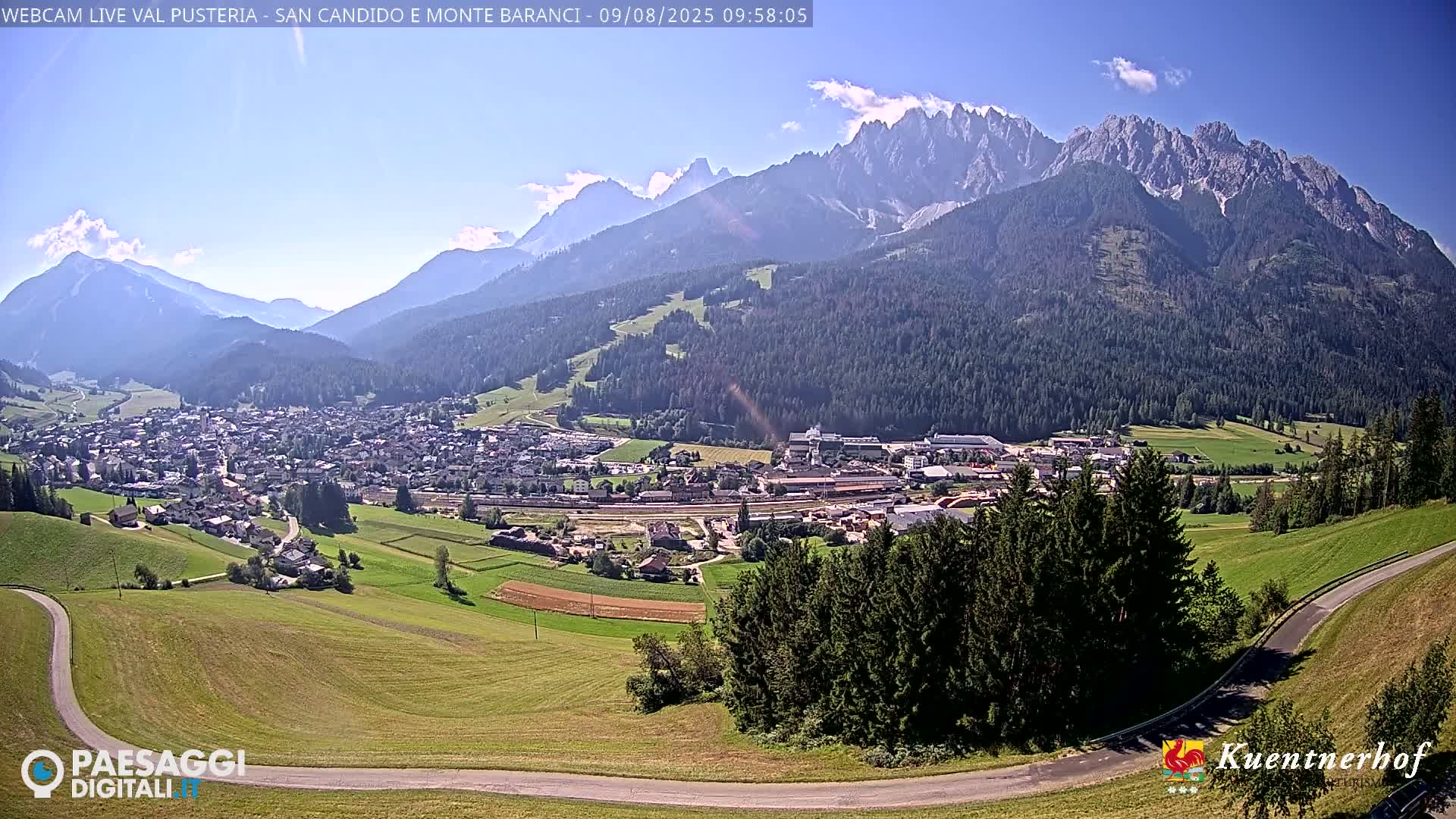 A sunny day reveals a valley town nestled beneath a towering, jagged mountain range.