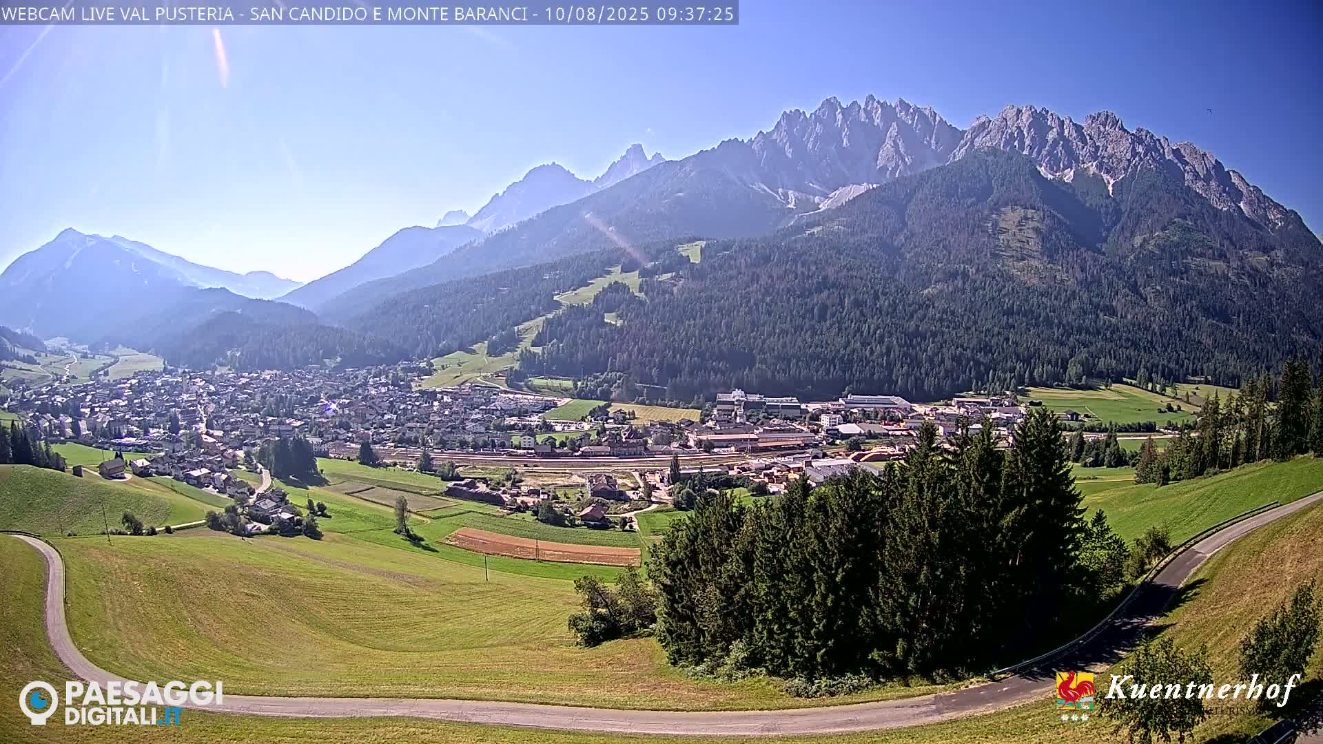 A sunny day reveals a valley town nestled below a large mountain range, with green fields and dark forests in between.