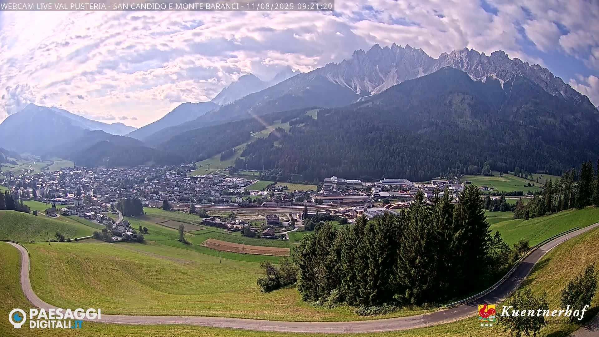 A mountain town nestled in a valley, under a partly cloudy sky, with a dramatic mountain range in the background.