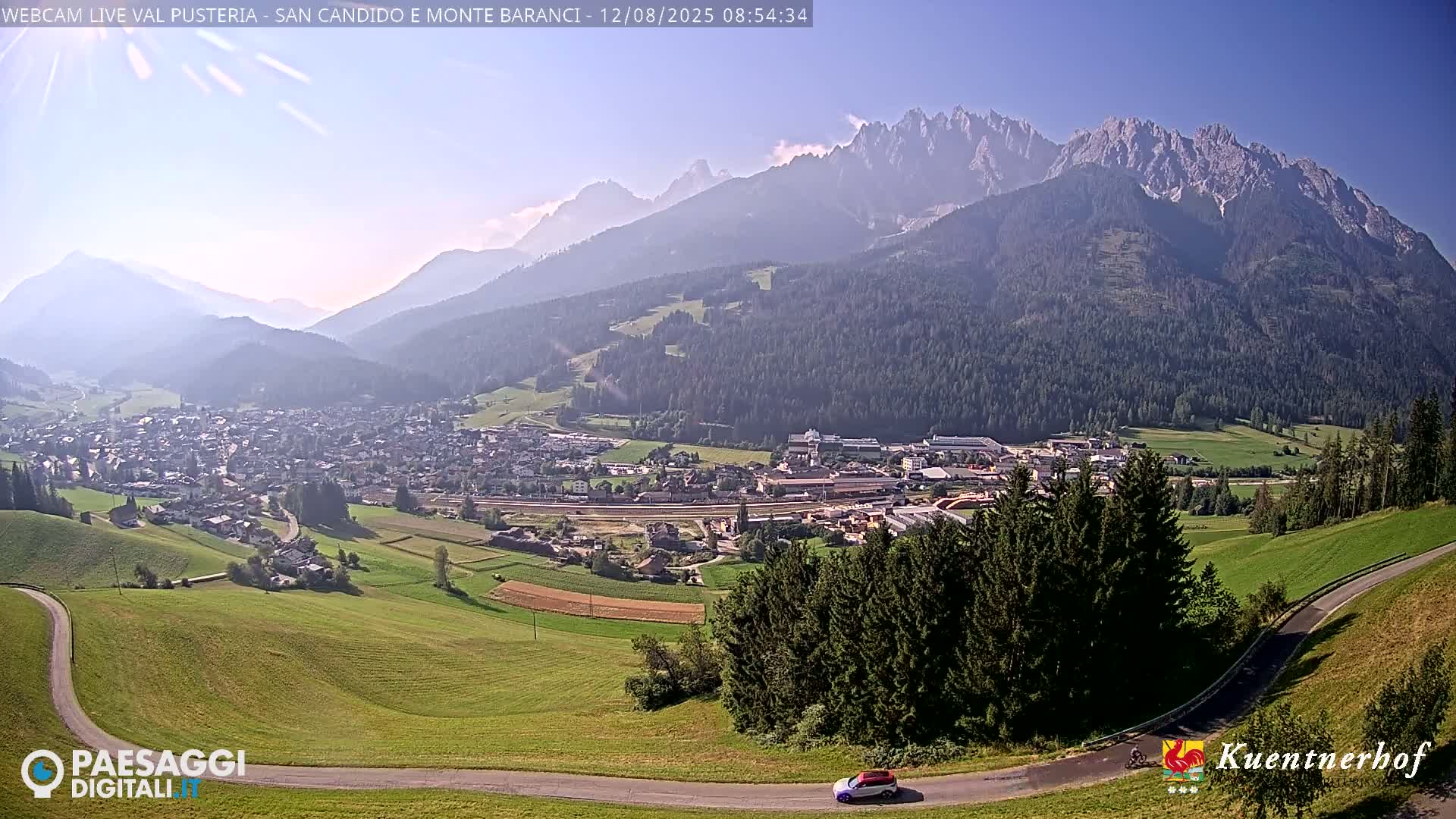 A sunny day reveals a valley town nestled beneath a hazy mountain range, with a winding road visible in the foreground.