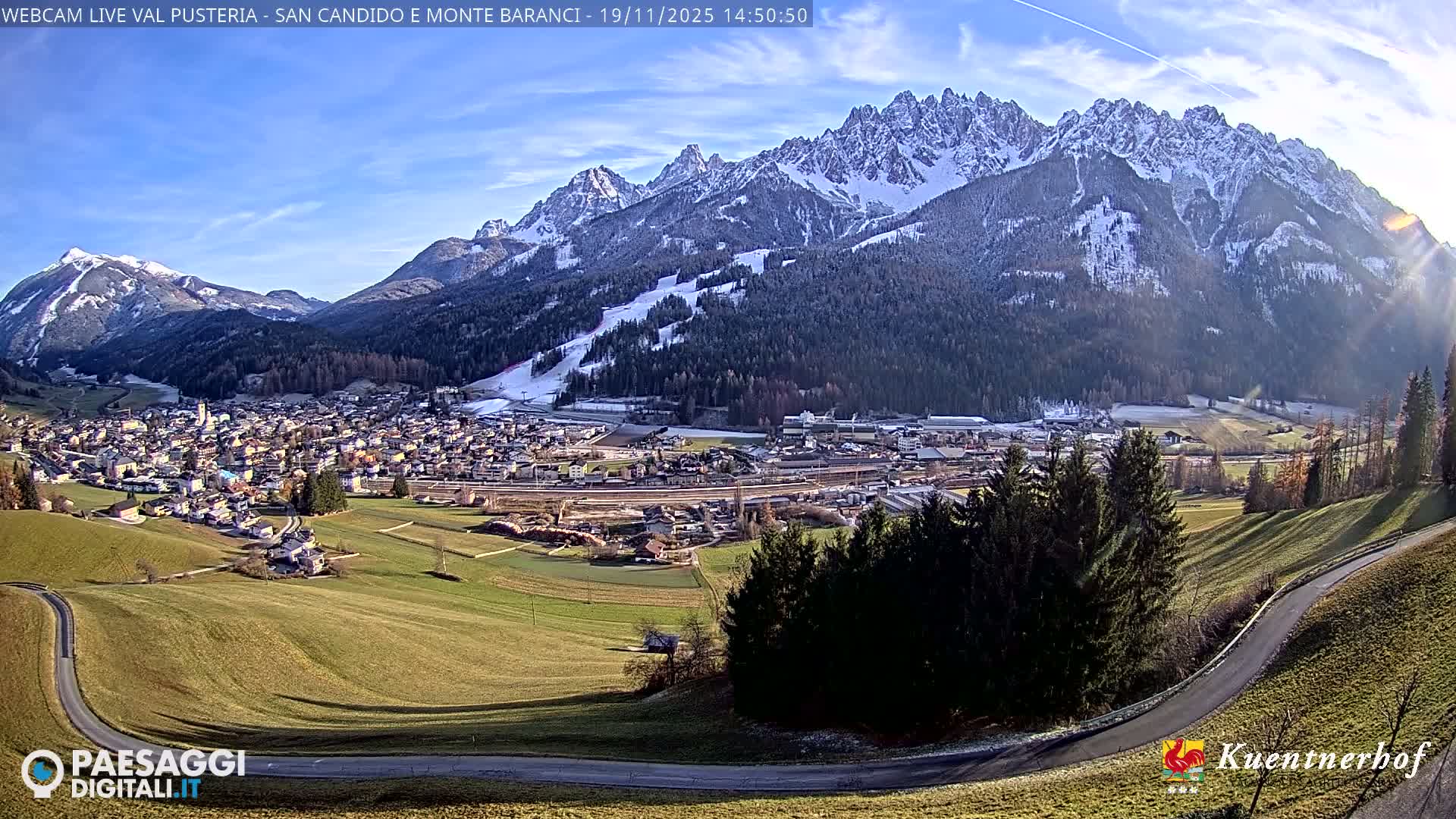 A sunny day reveals a valley town nestled beneath a hazy mountain range, with a winding road visible in the foreground.