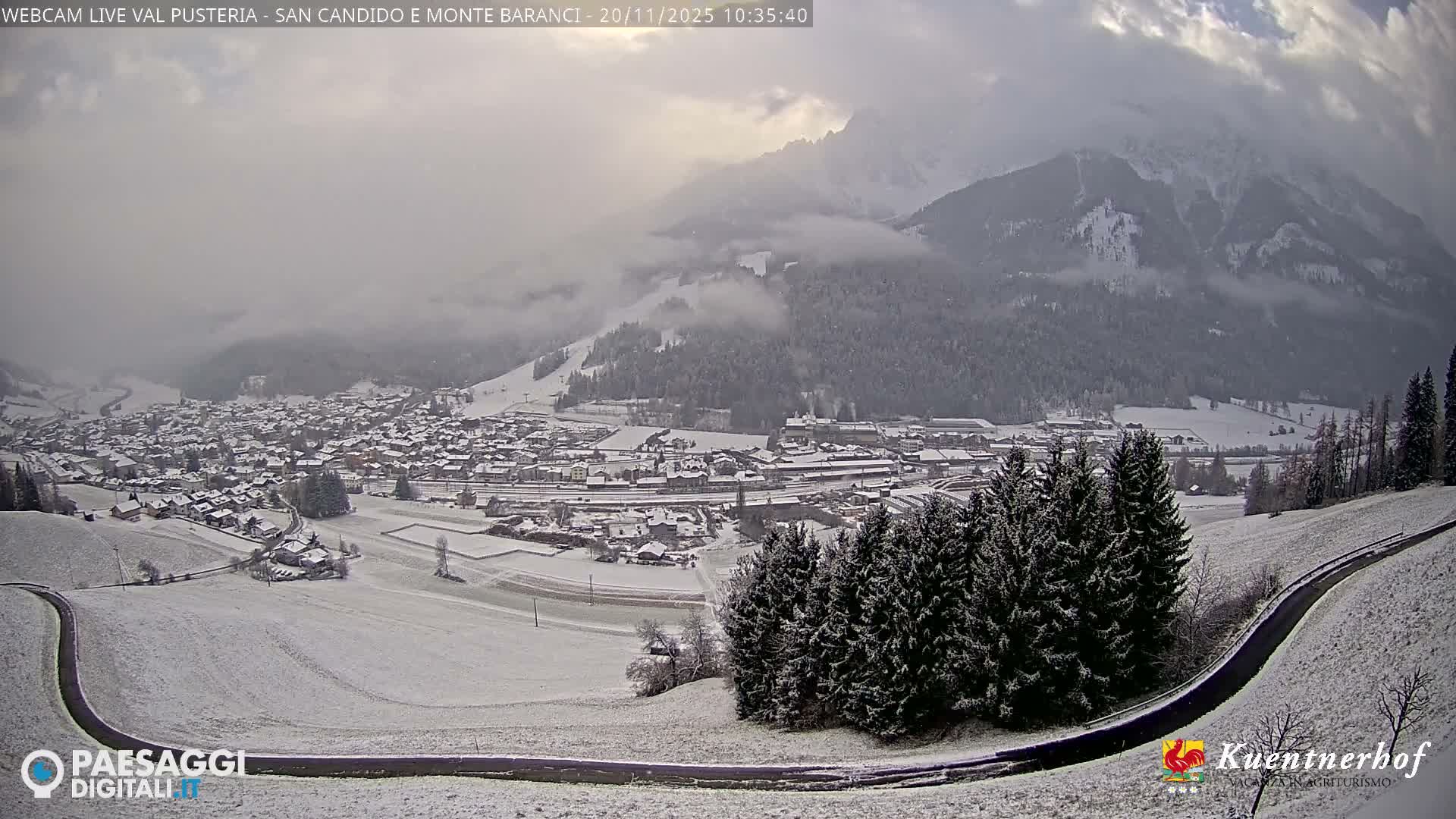 A panoramic view shows a snow-covered mountain village nestled in a valley with frosted evergreen trees and distant peaks under an overcast and hazy sky.