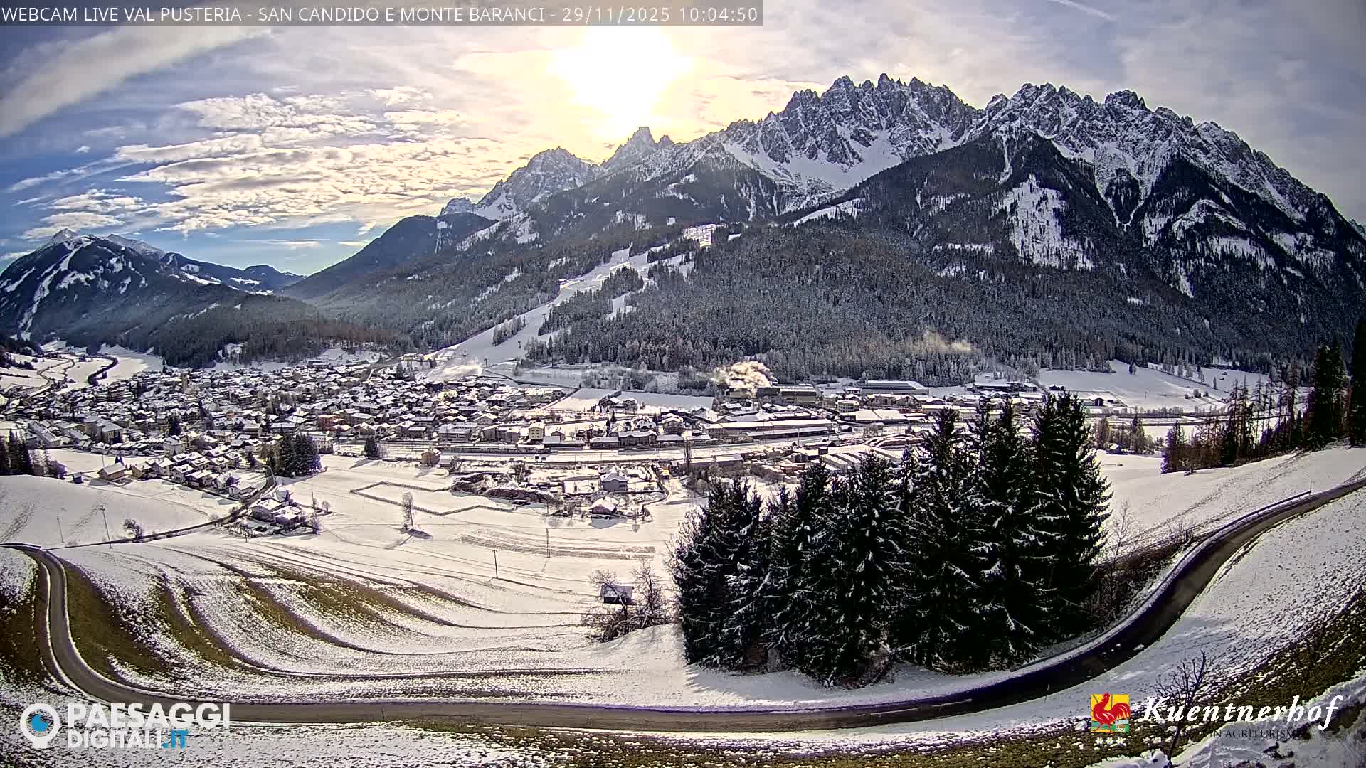 A scenic winter panorama captures a snow-dusted village set against majestic, snow-capped mountains under a partly cloudy sky with bright sunshine.