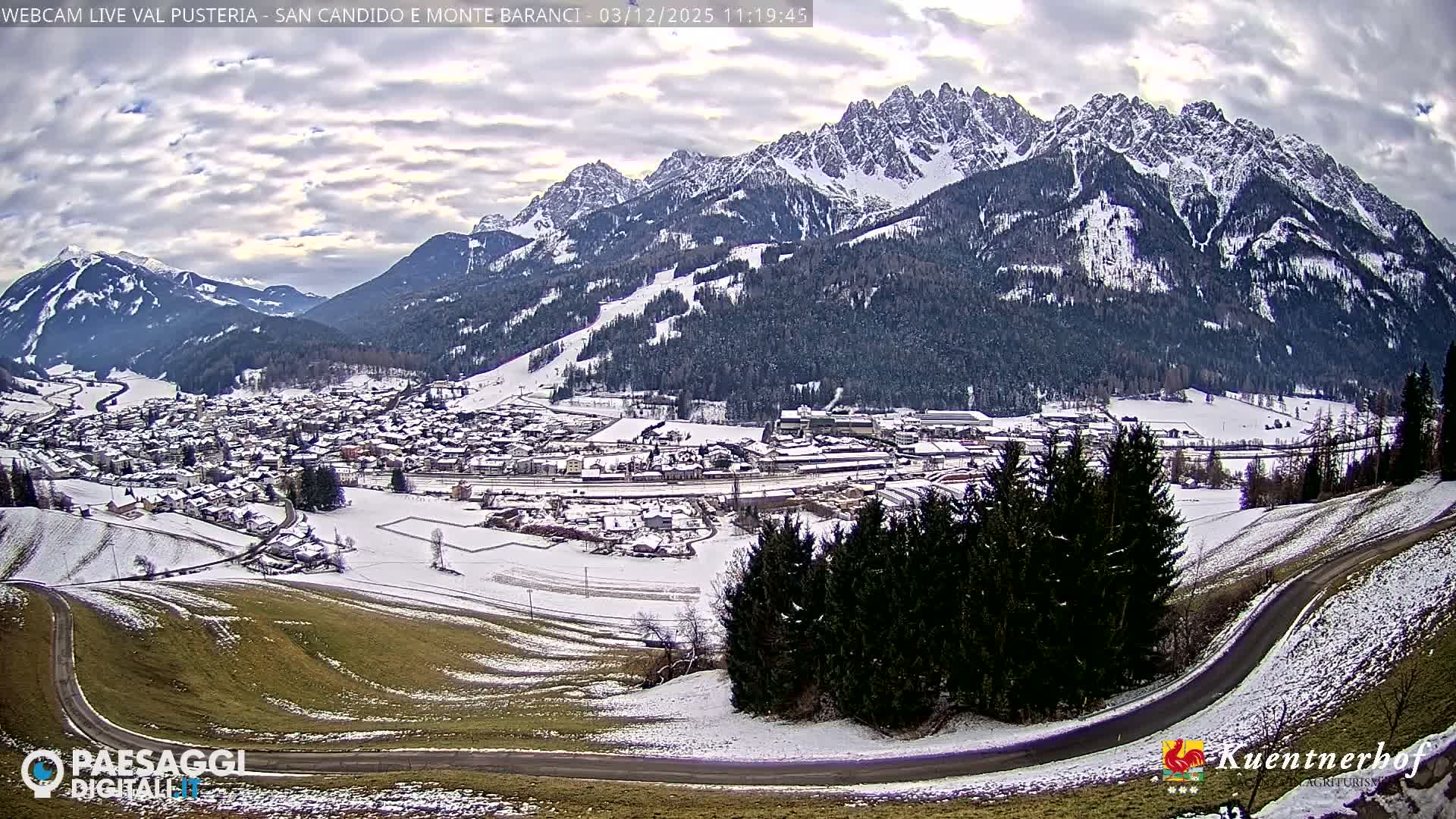 A vast winter landscape displays a snow-covered alpine village set in a valley, flanked by rugged, snow-capped mountains and evergreen forests under a cloudy sky, with a winding road and partial green slopes in the foreground.