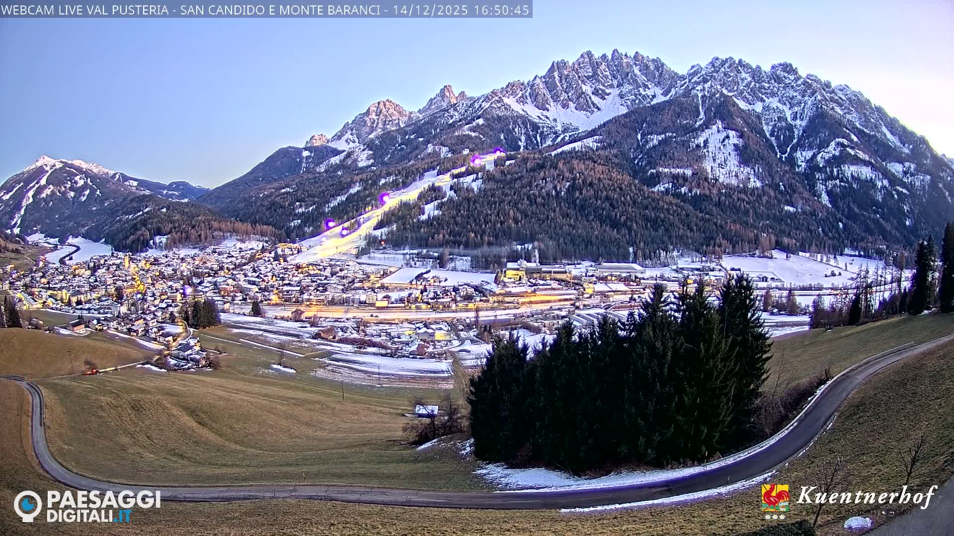 A vast winter landscape displays a snow-covered alpine village set in a valley, flanked by rugged, snow-capped mountains and evergreen forests under a cloudy sky, with a winding road and partial green slopes in the foreground.