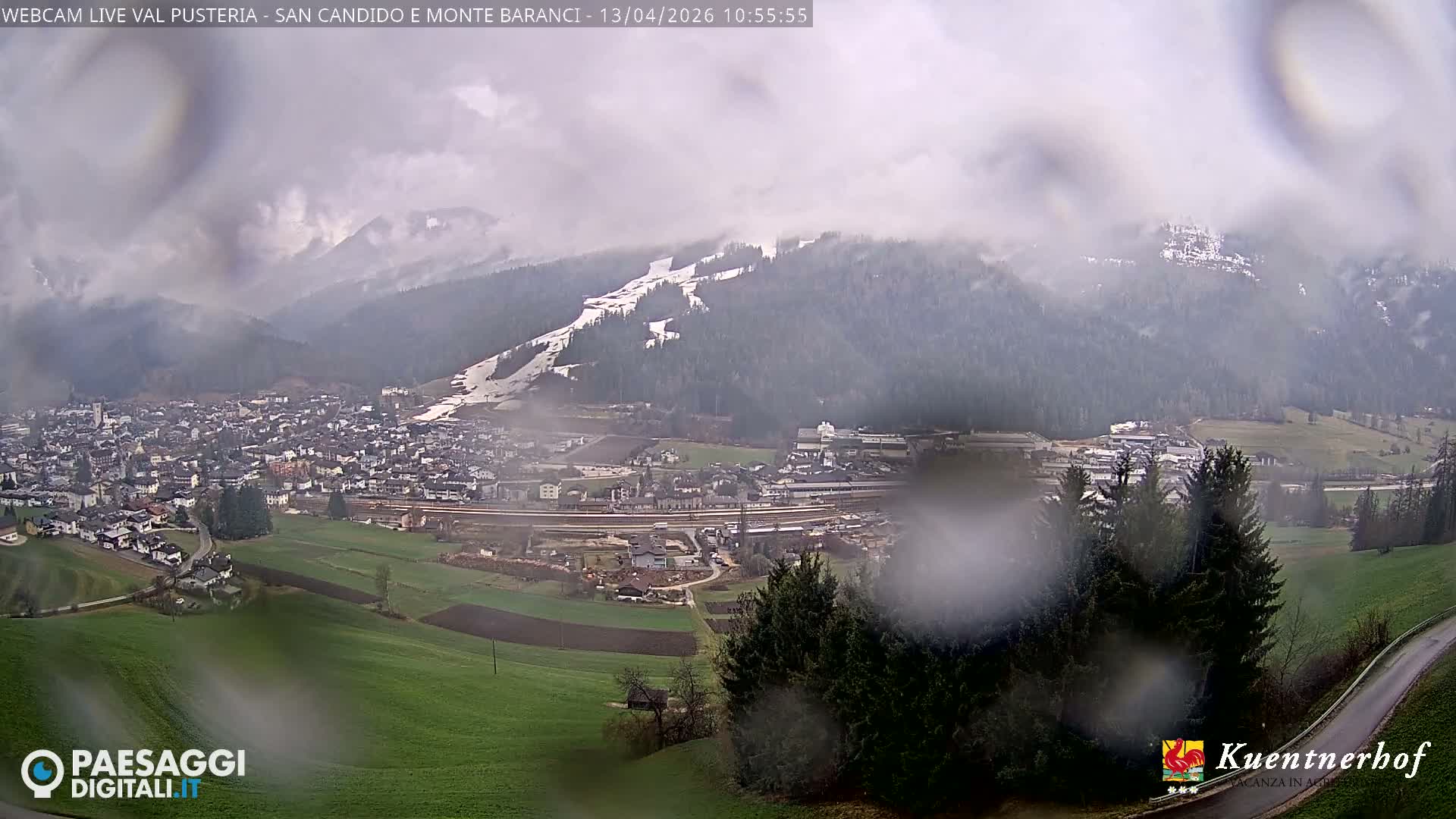 A vast winter landscape displays a snow-covered alpine village set in a valley, flanked by rugged, snow-capped mountains and evergreen forests under a cloudy sky, with a winding road and partial green slopes in the foreground.