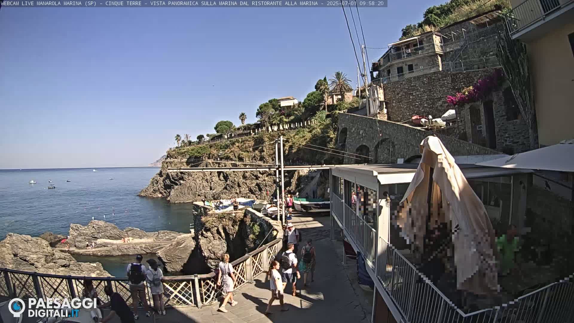 A sunny seaside village scene shows people walking along a path near a rocky coastline with boats in the calm water.