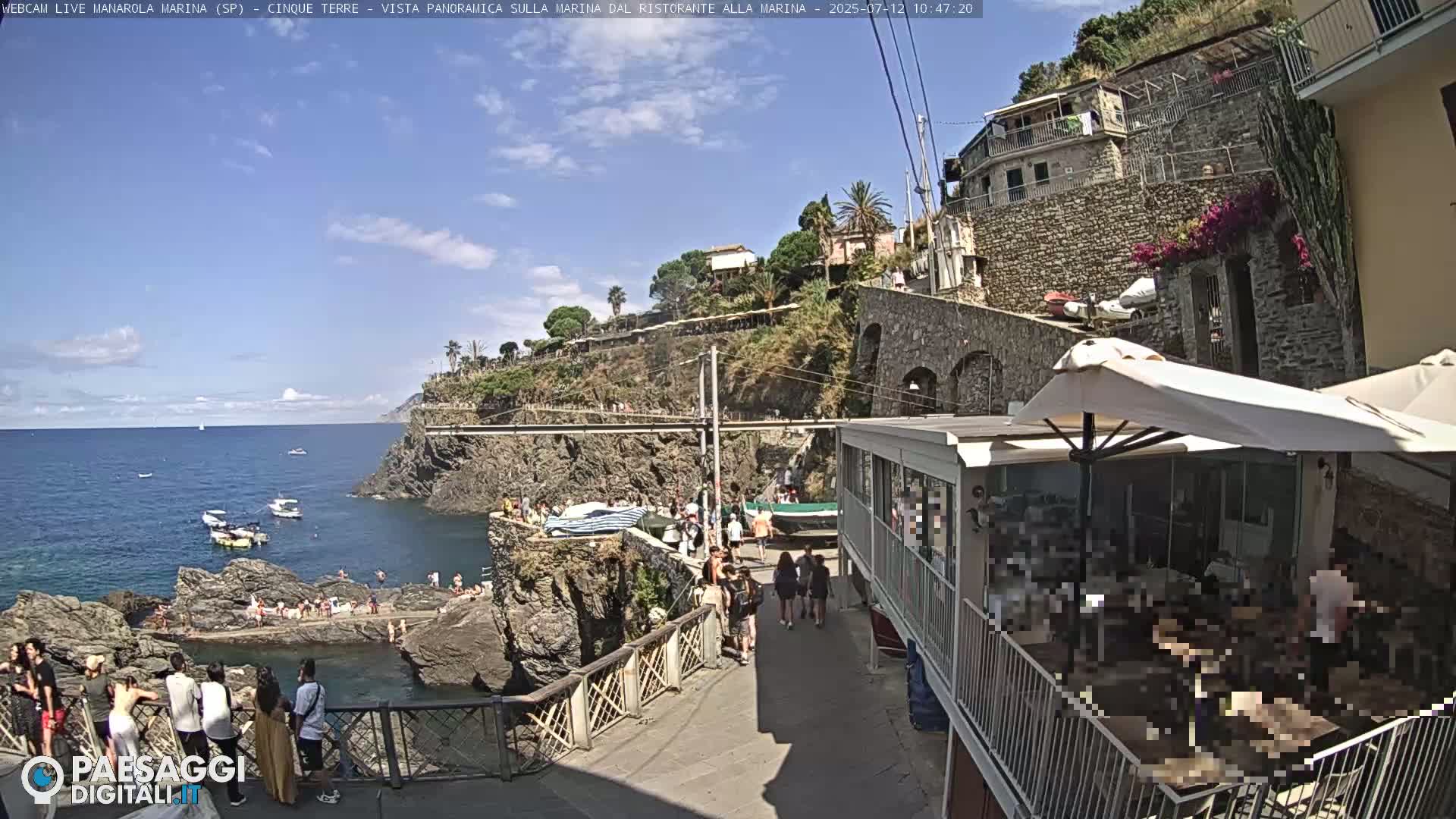 A sunny seaside village scene shows people walking along a path near a rocky coastline with boats in the calm water.