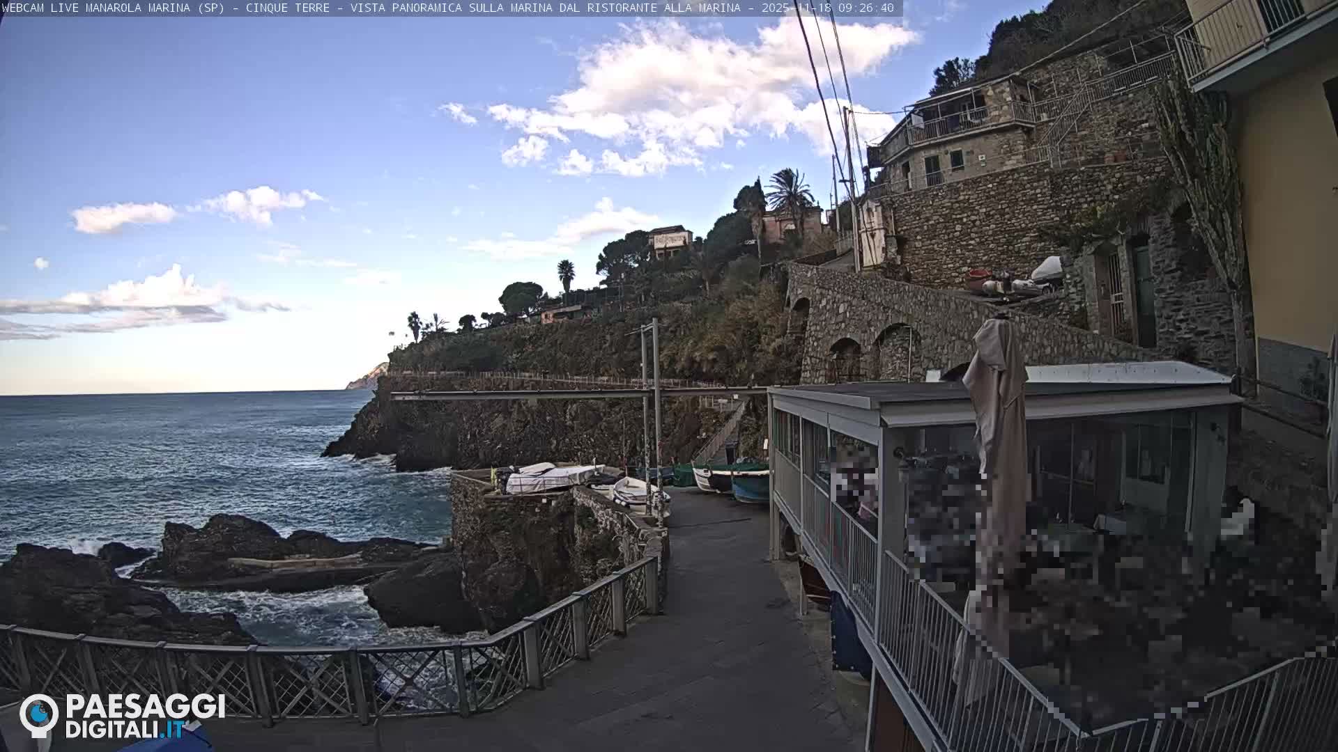 A scenic daytime view depicts a rocky coastal town with blue-green waters crashing against the shore, several small boats pulled ashore along a waterfront path, and traditional stone buildings climbing a tree-lined hillside under a partly cloudy sky.