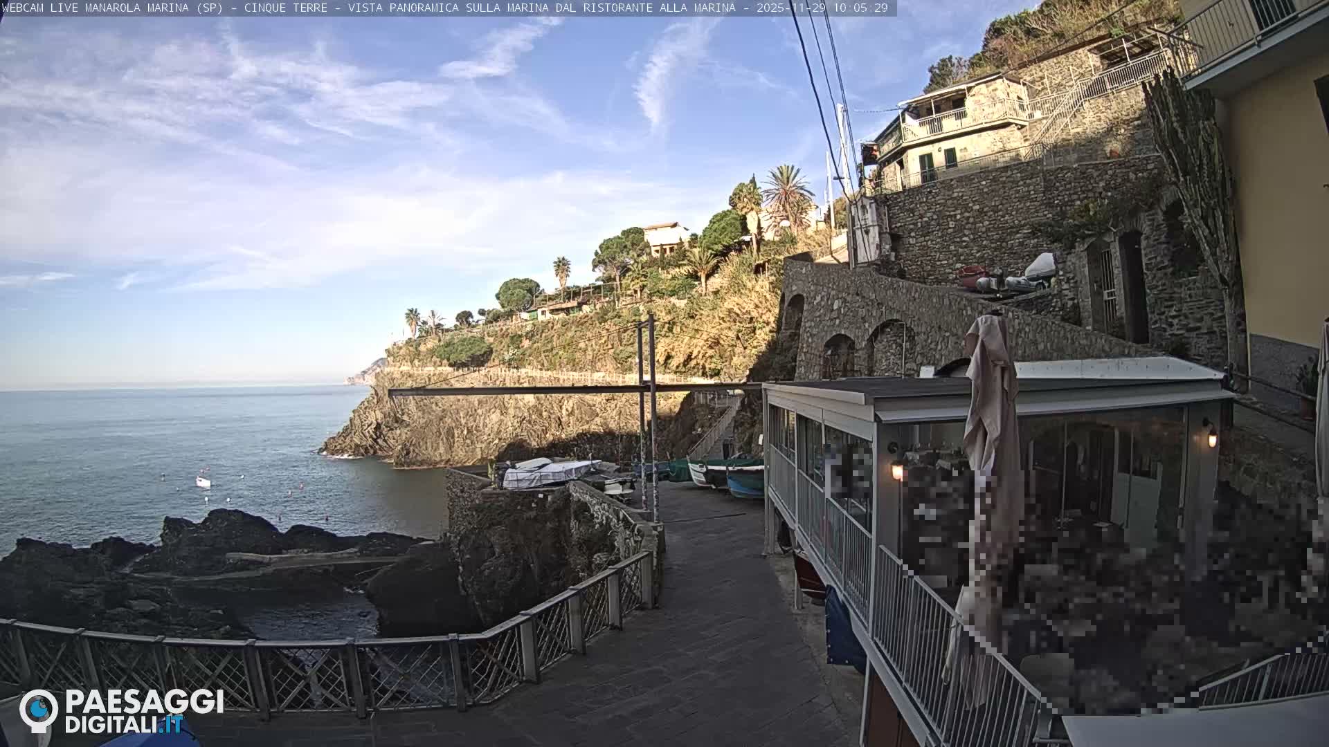 A sunny day illuminates a picturesque coastal village, revealing traditional stone buildings clinging to steep, vegetated cliffs overlooking a rocky marina with parked boats and a seaside restaurant, all under a clear blue sky with sparse clouds.