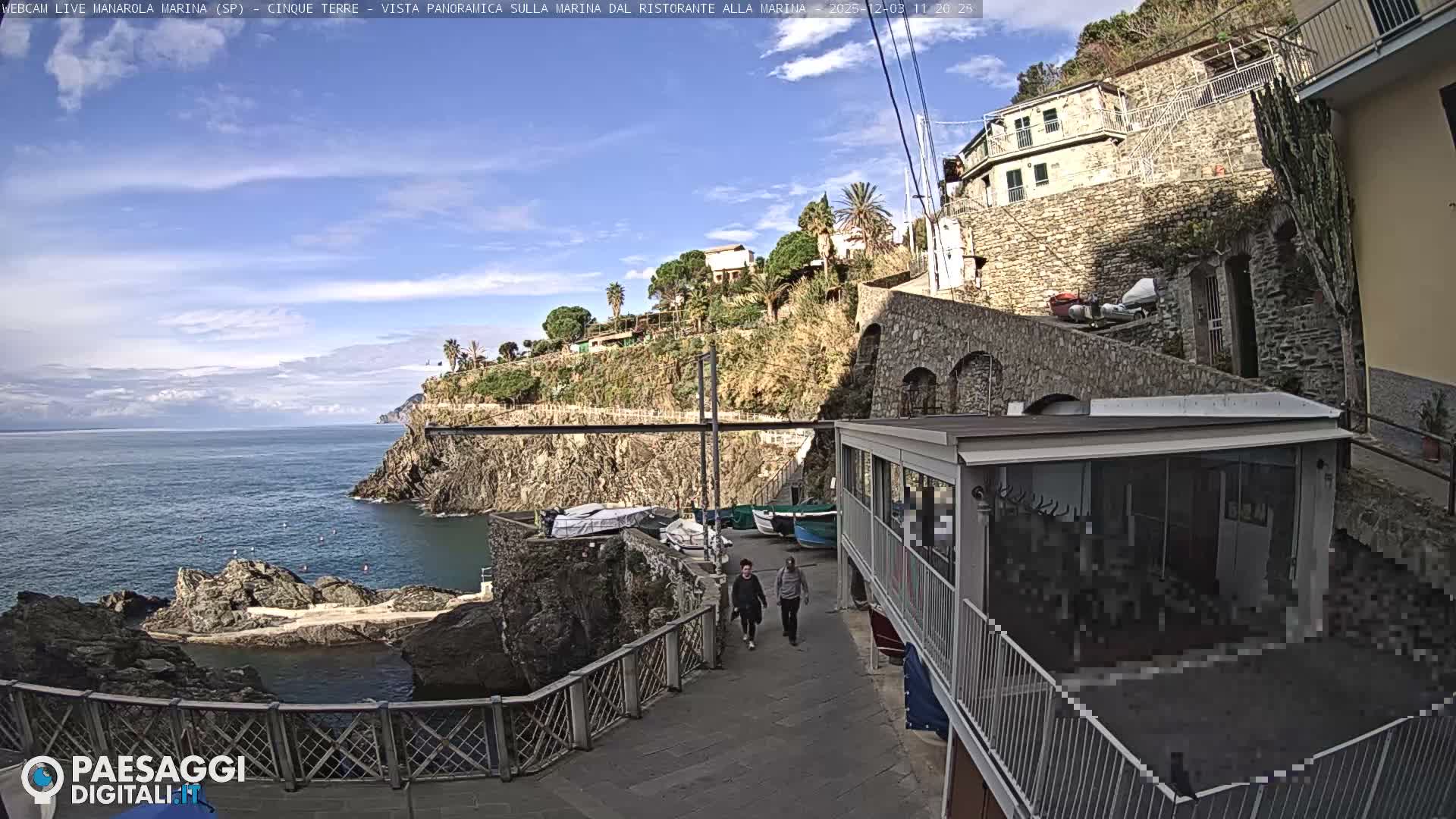 Under a sunny, partly cloudy sky, two people stroll along a waterfront path past boats and a cafe, with the blue sea and rocky cliffs on the left, and a hillside village featuring stone buildings and lush vegetation on the right.