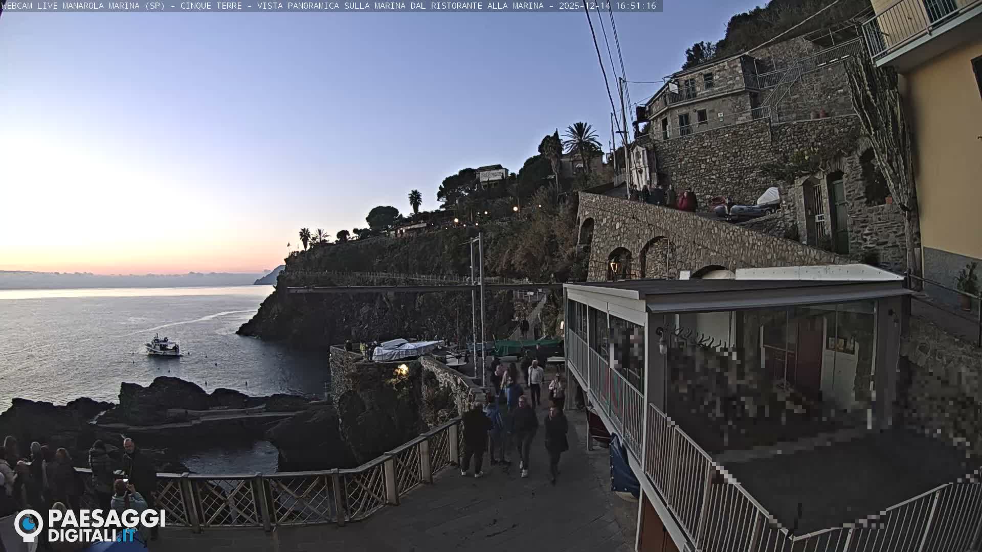 Under a sunny, partly cloudy sky, two people stroll along a waterfront path past boats and a cafe, with the blue sea and rocky cliffs on the left, and a hillside village featuring stone buildings and lush vegetation on the right.