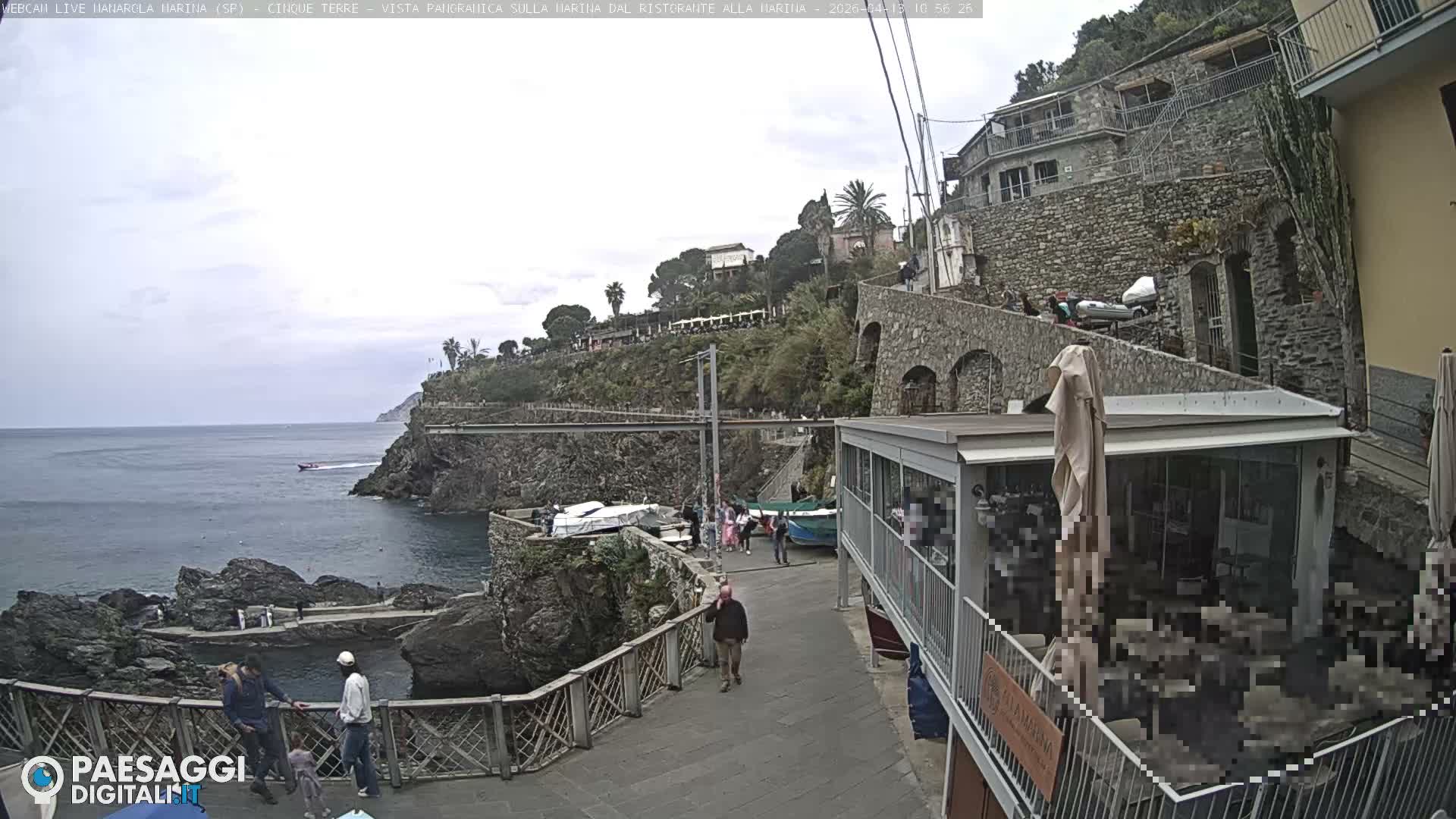 Under a sunny, partly cloudy sky, two people stroll along a waterfront path past boats and a cafe, with the blue sea and rocky cliffs on the left, and a hillside village featuring stone buildings and lush vegetation on the right.