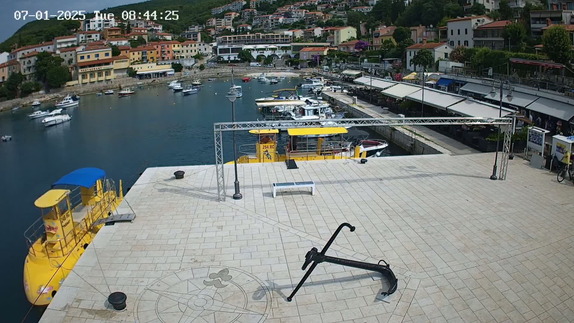 A yellow submarine and several boats are docked in a harbor next to a paved area with an anchor and buildings on a hillside in the background under a sunny sky.
