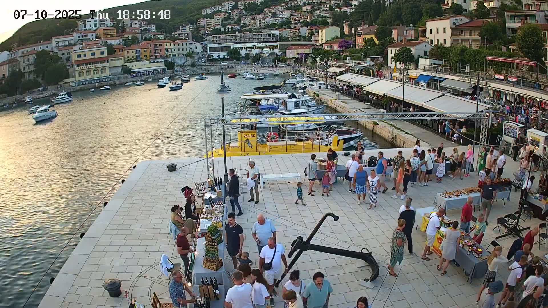 A harbor town at sunset with many people gathered at an outdoor market on a pier, numerous boats docked in the water, and buildings on a hillside in the background.