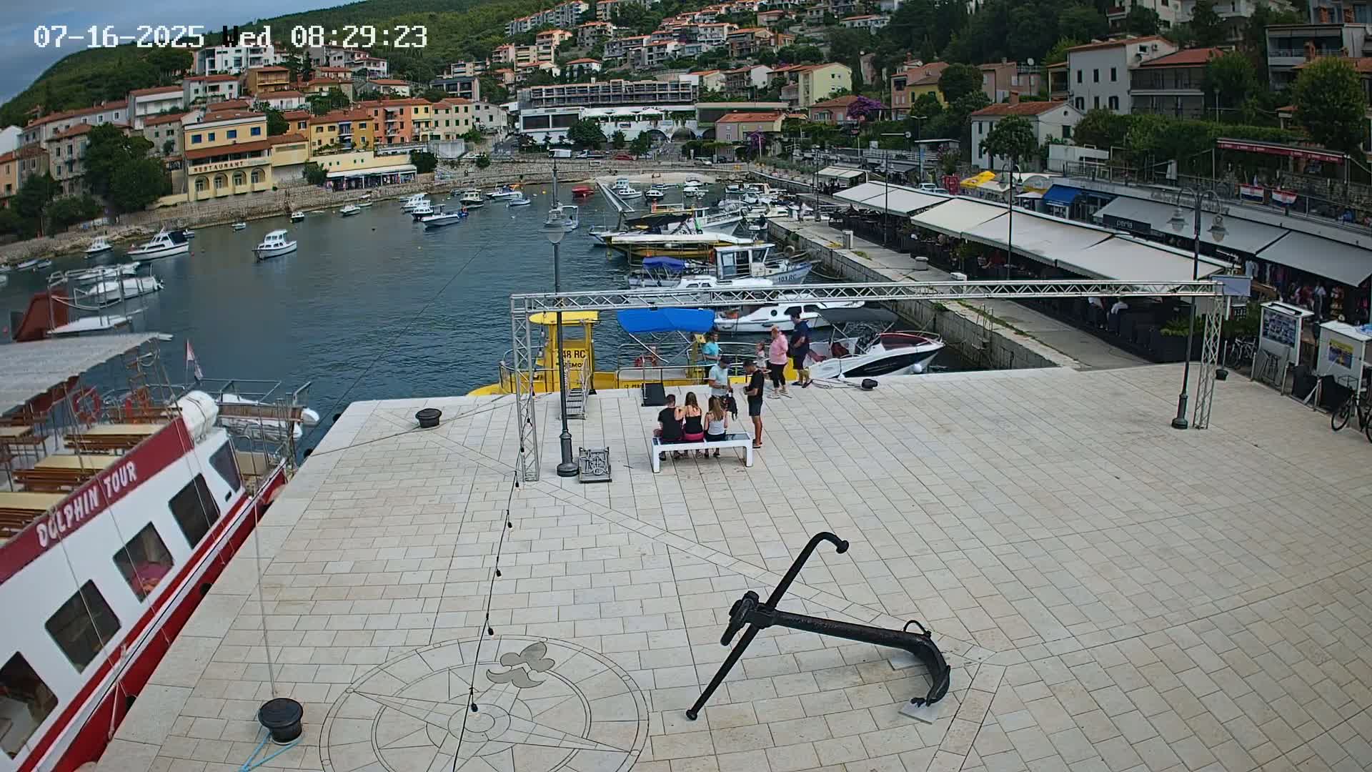 A harbor scene on a sunny day shows a paved area with a large anchor, several people sitting at a table, and numerous boats moored in the calm water; buildings and trees are visible in the background.