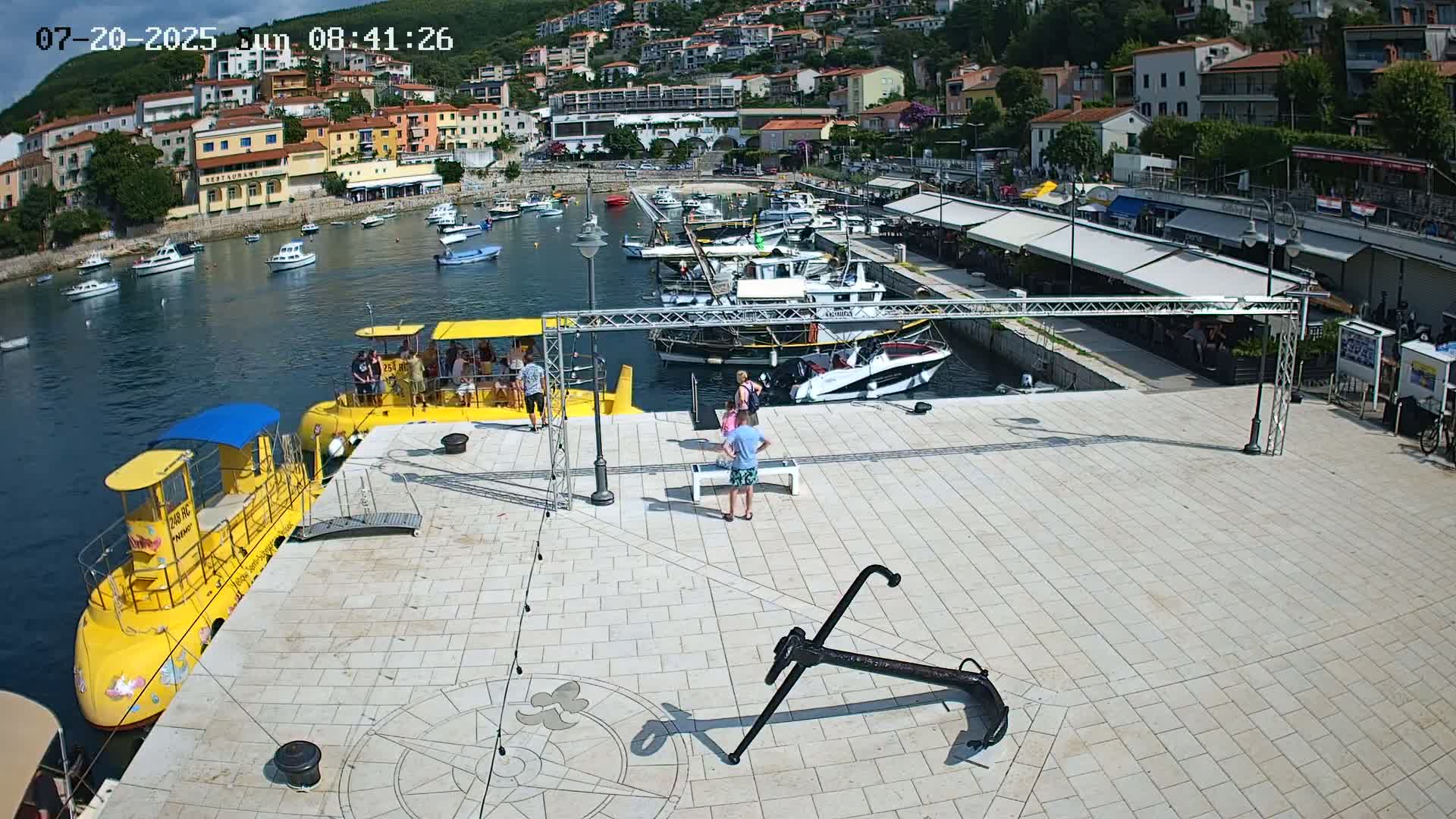 A harbor with numerous boats, a yellow submarine, and several people on a paved area, under a partly cloudy sky.