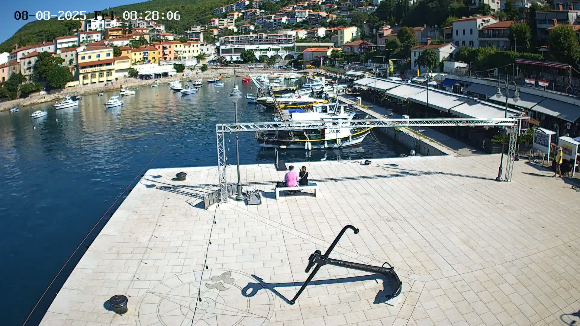 A couple sits on a bench overlooking a harbor filled with boats on a sunny day, with a large anchor and compass rose on the paved surface in the foreground.