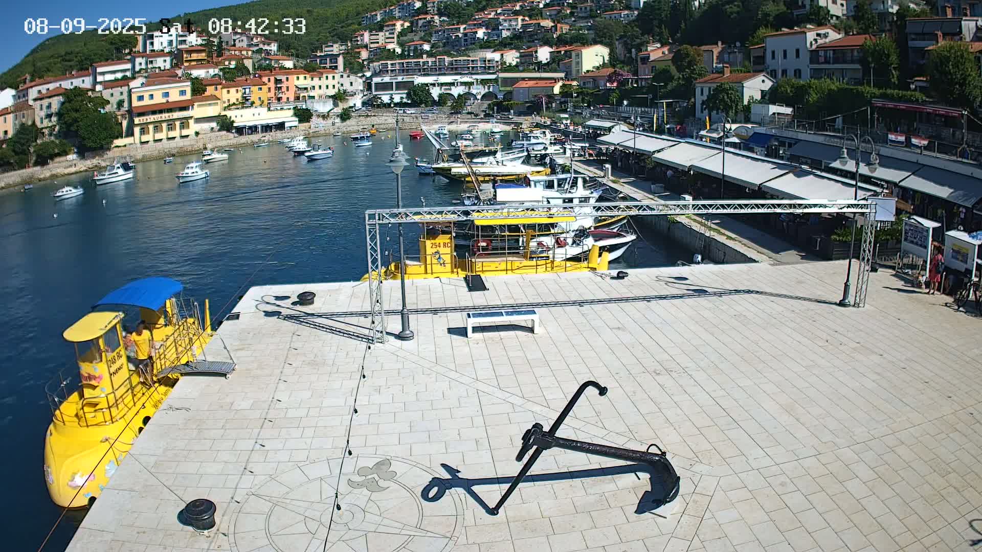 A sunny harbor scene shows a yellow submarine docked near a large anchor on a paved area, with numerous boats in the water and buildings on a hillside in the background.
