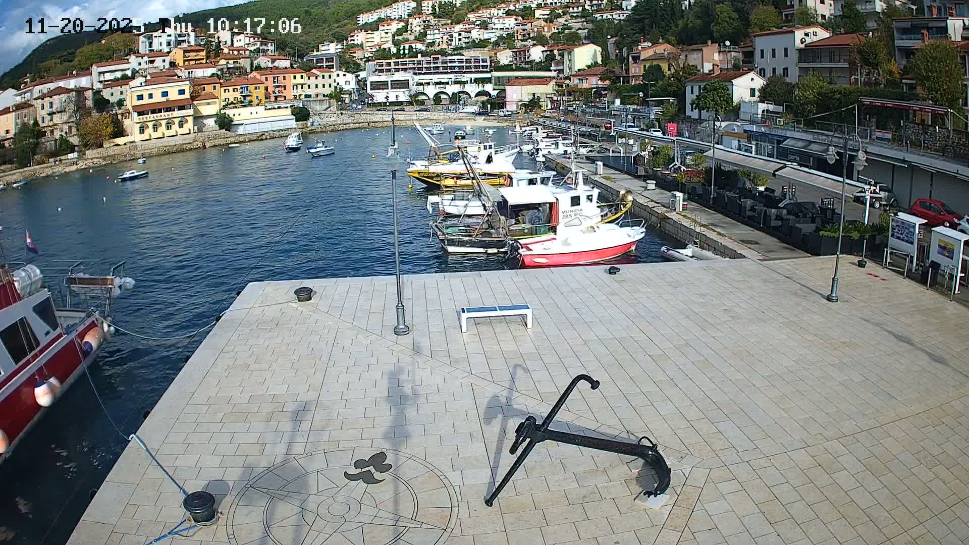 On a clear and sunny day, a bustling harbor is depicted with numerous boats docked in calm blue water, set against a backdrop of a colorful hillside town with buildings ascending the green slopes.