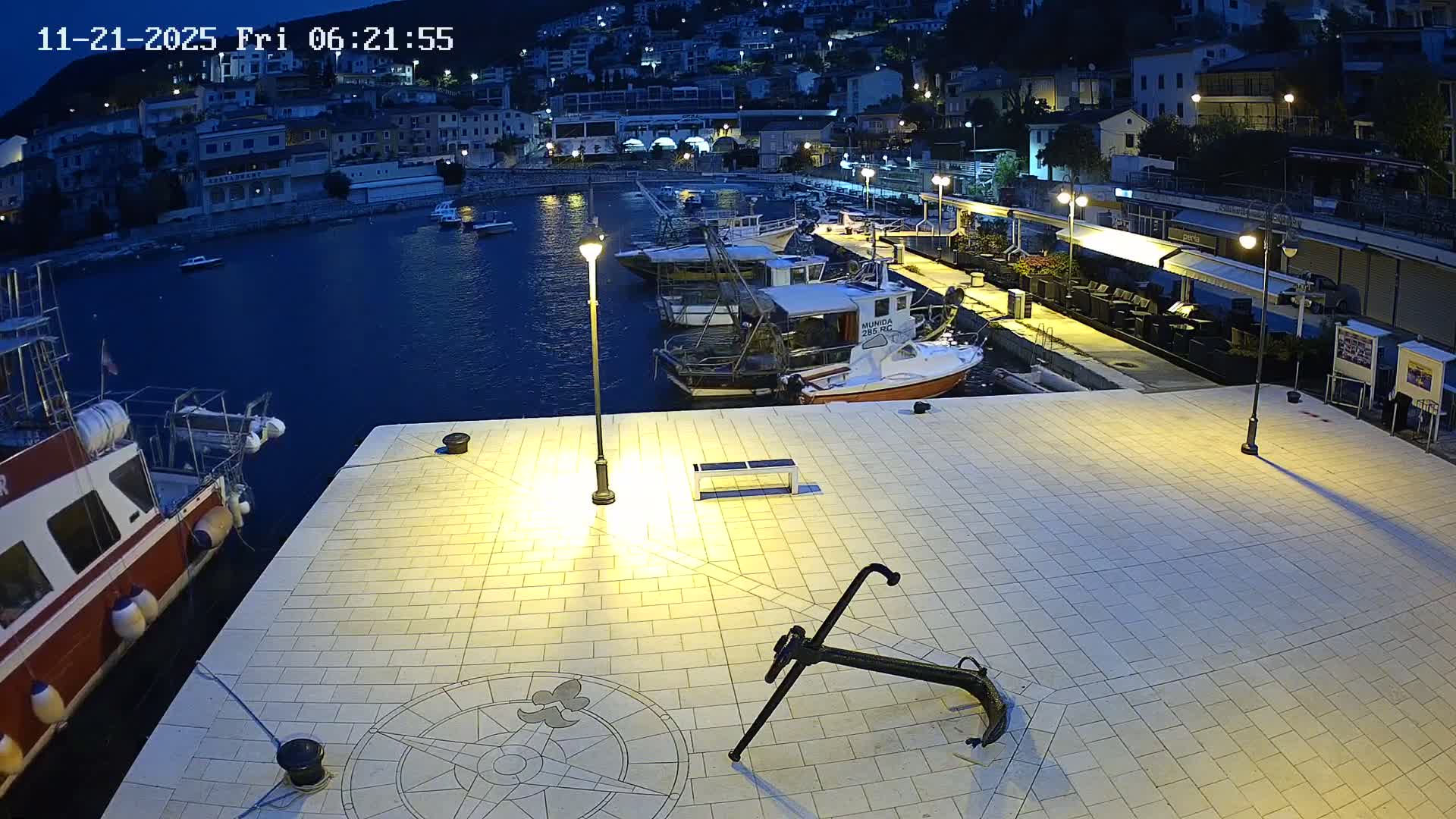 On a clear and sunny day, a bustling harbor is depicted with numerous boats docked in calm blue water, set against a backdrop of a colorful hillside town with buildings ascending the green slopes.