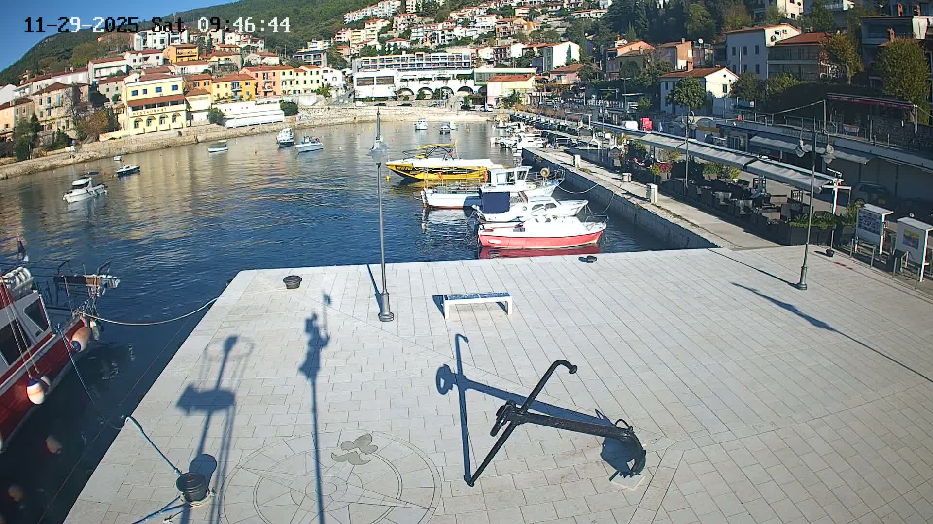 A sunny harbor scene features numerous boats moored in blue water, a paved promenade with a large anchor sculpture and outdoor seating areas, all set against a backdrop of a colorful town built into a lush, green hillside.
