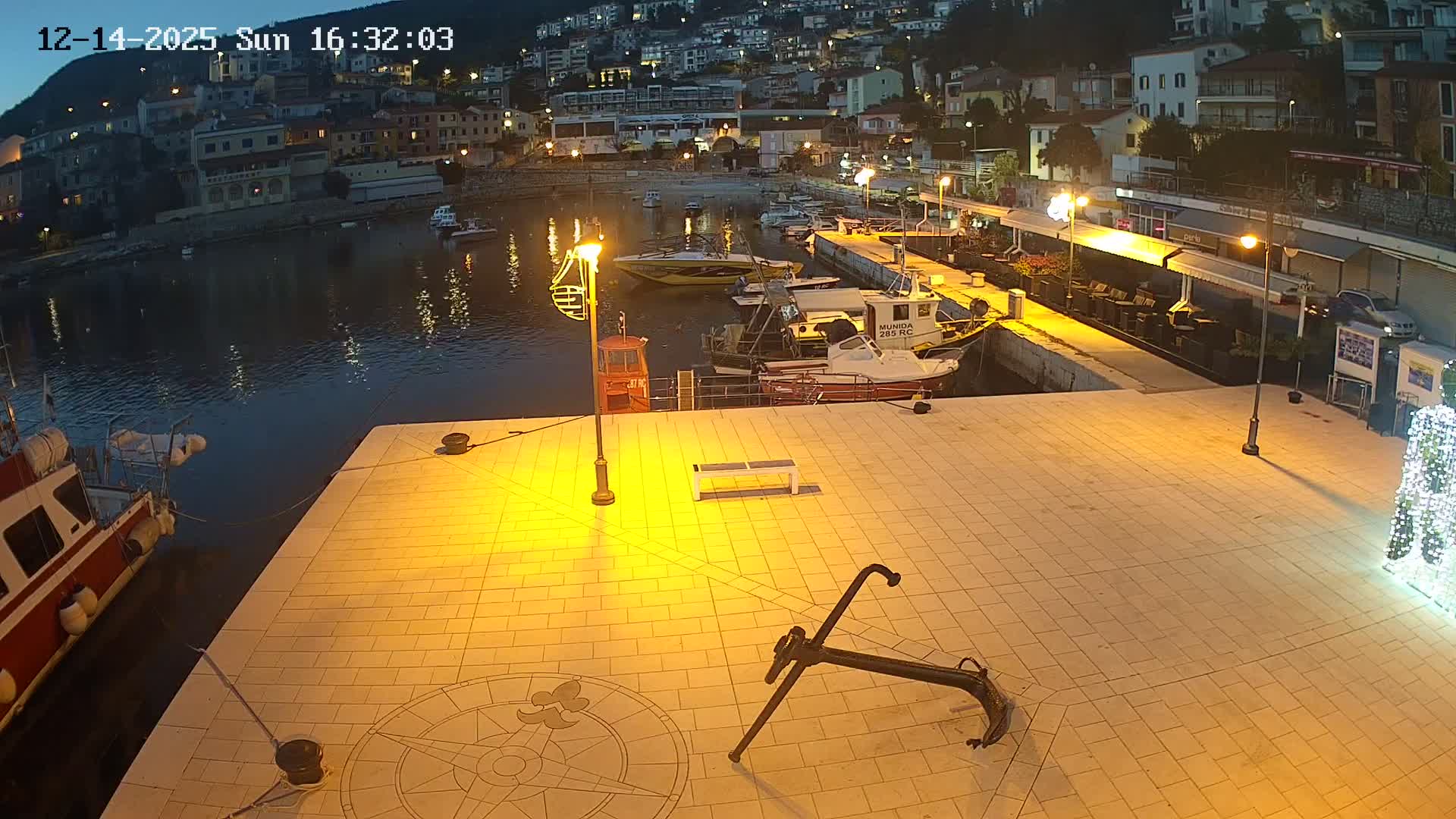 A sunny harbor scene features numerous boats moored in blue water, a paved promenade with a large anchor sculpture and outdoor seating areas, all set against a backdrop of a colorful town built into a lush, green hillside.