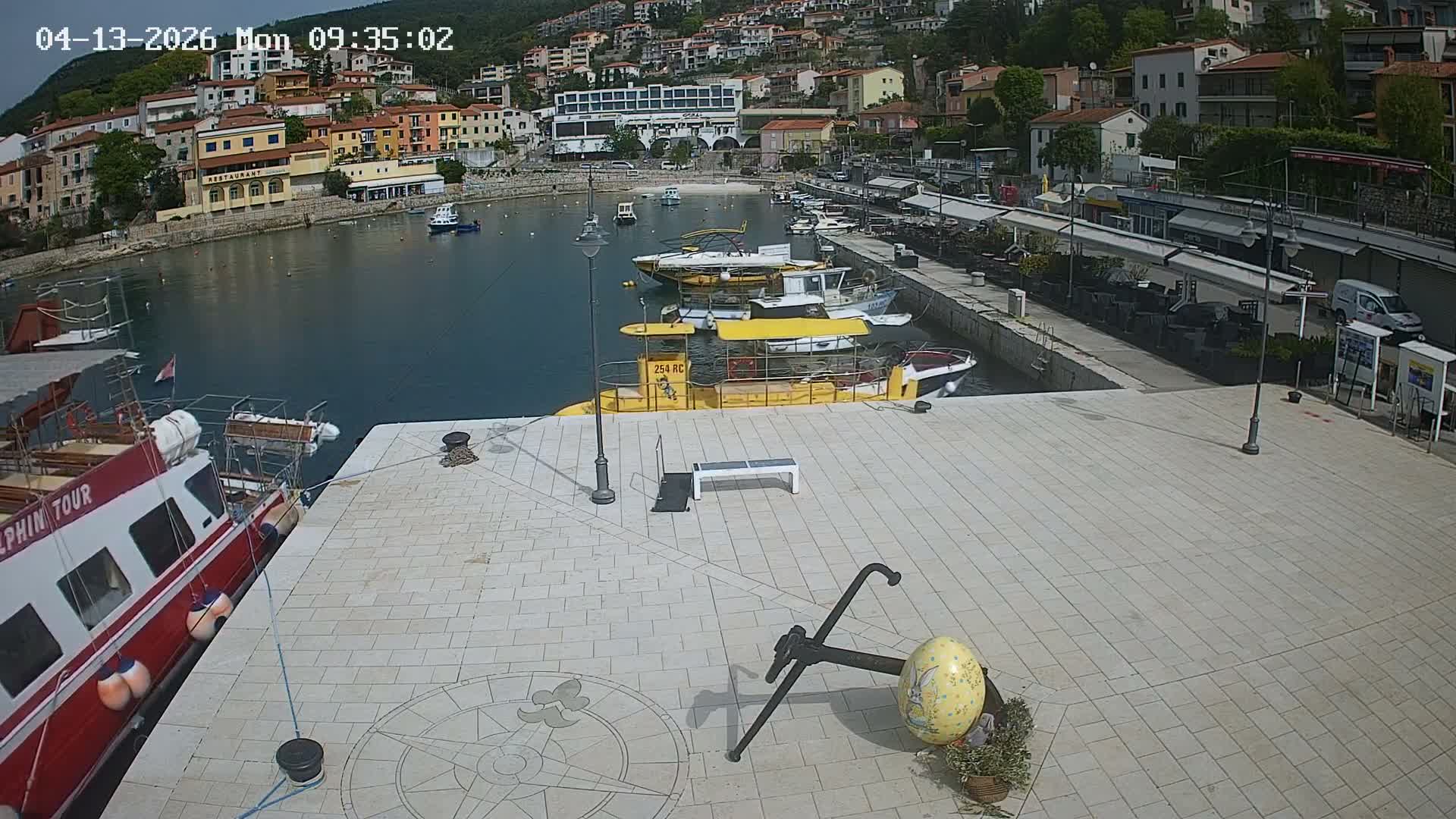 A sunny harbor scene features numerous boats moored in blue water, a paved promenade with a large anchor sculpture and outdoor seating areas, all set against a backdrop of a colorful town built into a lush, green hillside.
