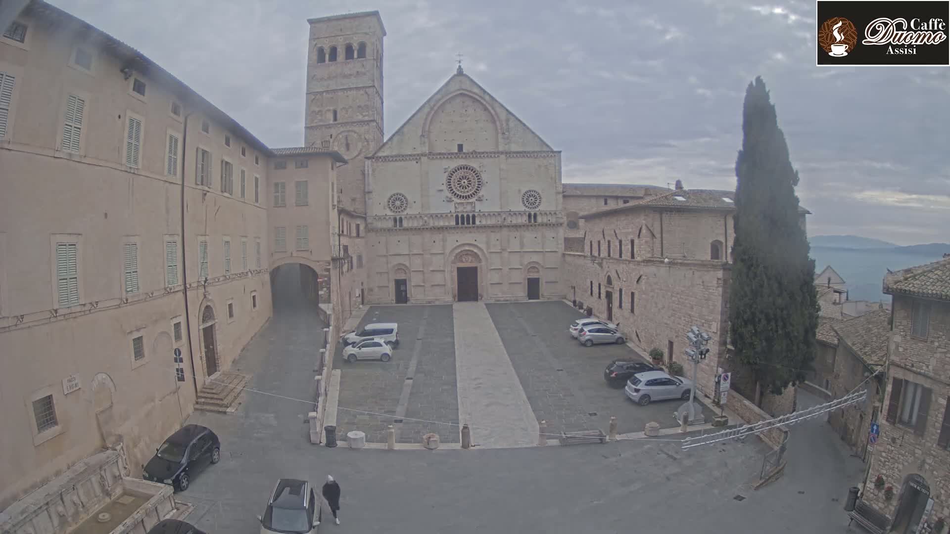 A large stone church stands in a plaza with several cars parked, all under an overcast sky.