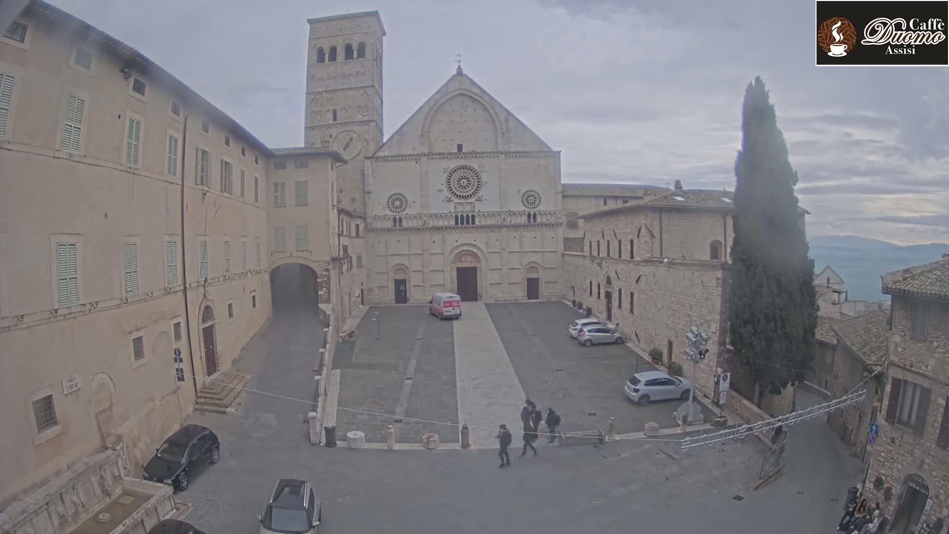 A large stone cathedral stands in a town square on an overcast day, surrounded by other stone buildings and a few parked cars.