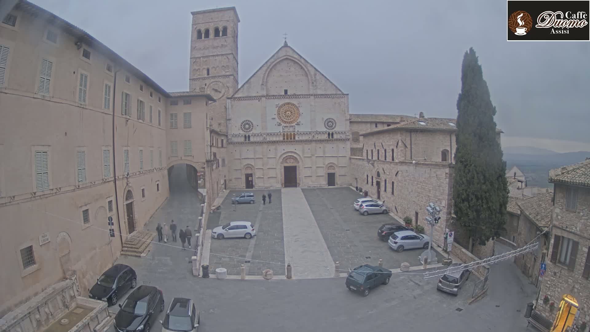 A large stone cathedral faces a plaza with several parked cars, surrounded by older stone buildings under an overcast sky.